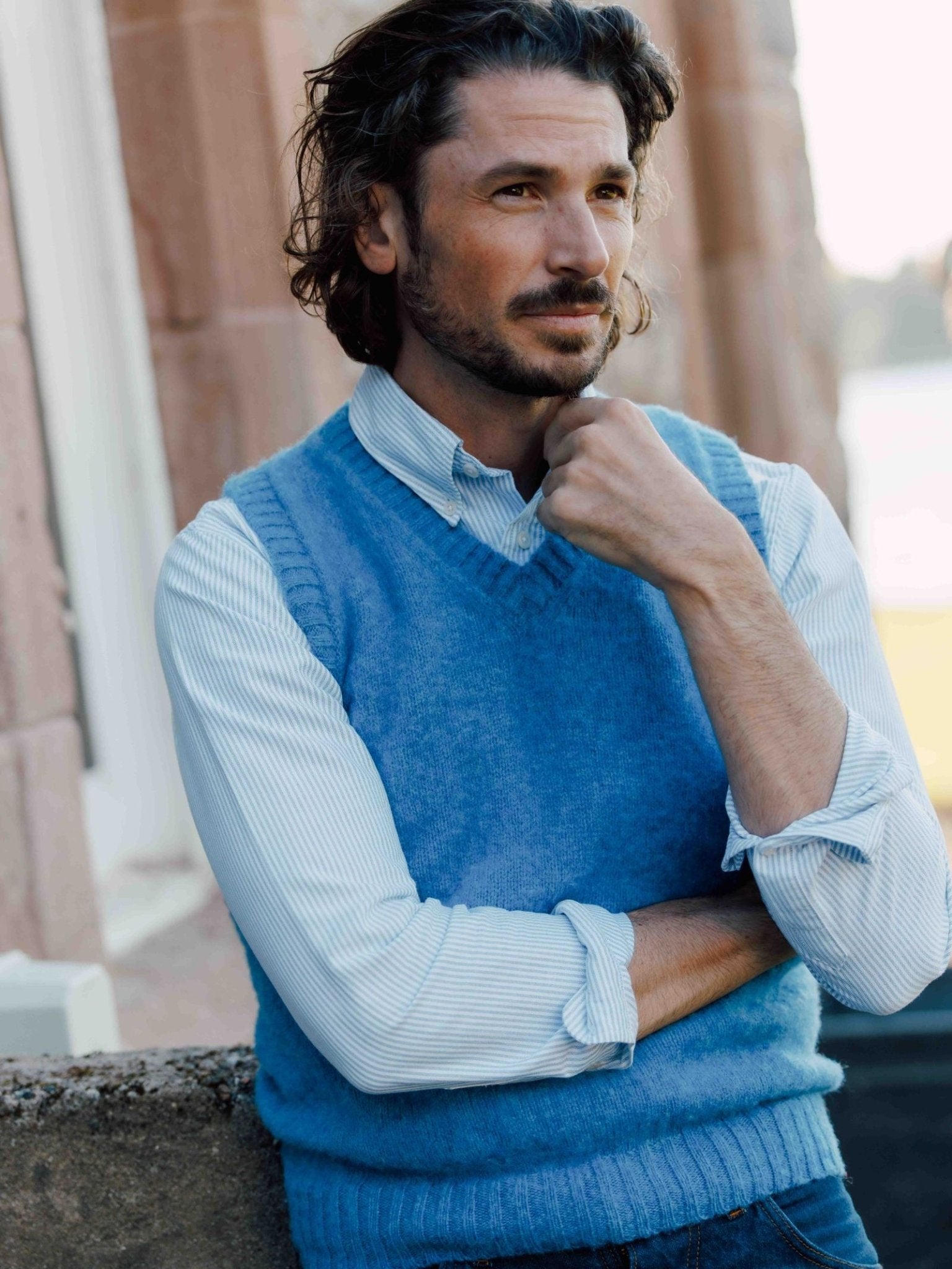 A man with wavy brown hair and a beard, dressed in a Campbells of Beauly Oxford Shirt with a light blue sweater vest, stands outdoors by a stone building, resting his chin thoughtfully on his hand.