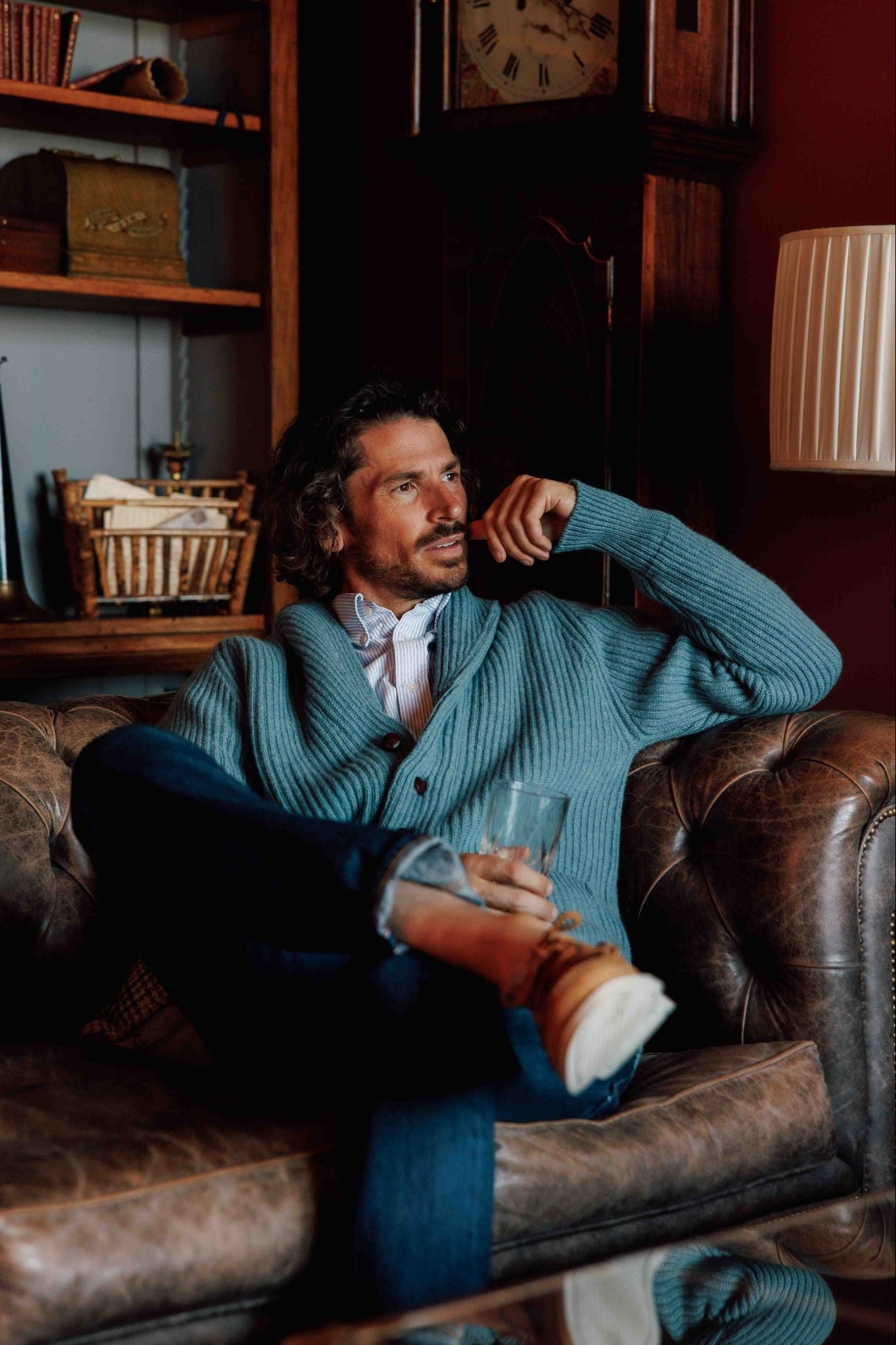 A man with wavy hair, wearing the Campbell's of Beauly Cashmere Shawl Collar Cardigan in light blue and jeans, sits relaxed on a brown leather couch with a glass in hand, gazing thoughtfully to the side. Shelves and a large clock are visible behind him.