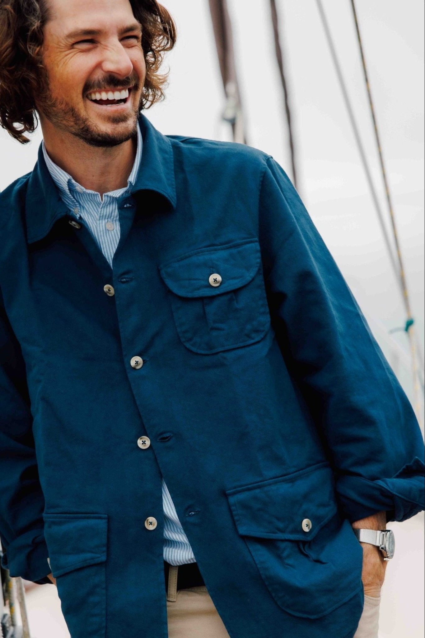 A smiling man with curly brown hair wears the Campbell's of Beauly Auld Stock Field Chore Jacket, made in Europe from 100% cotton, over a striped shirt.