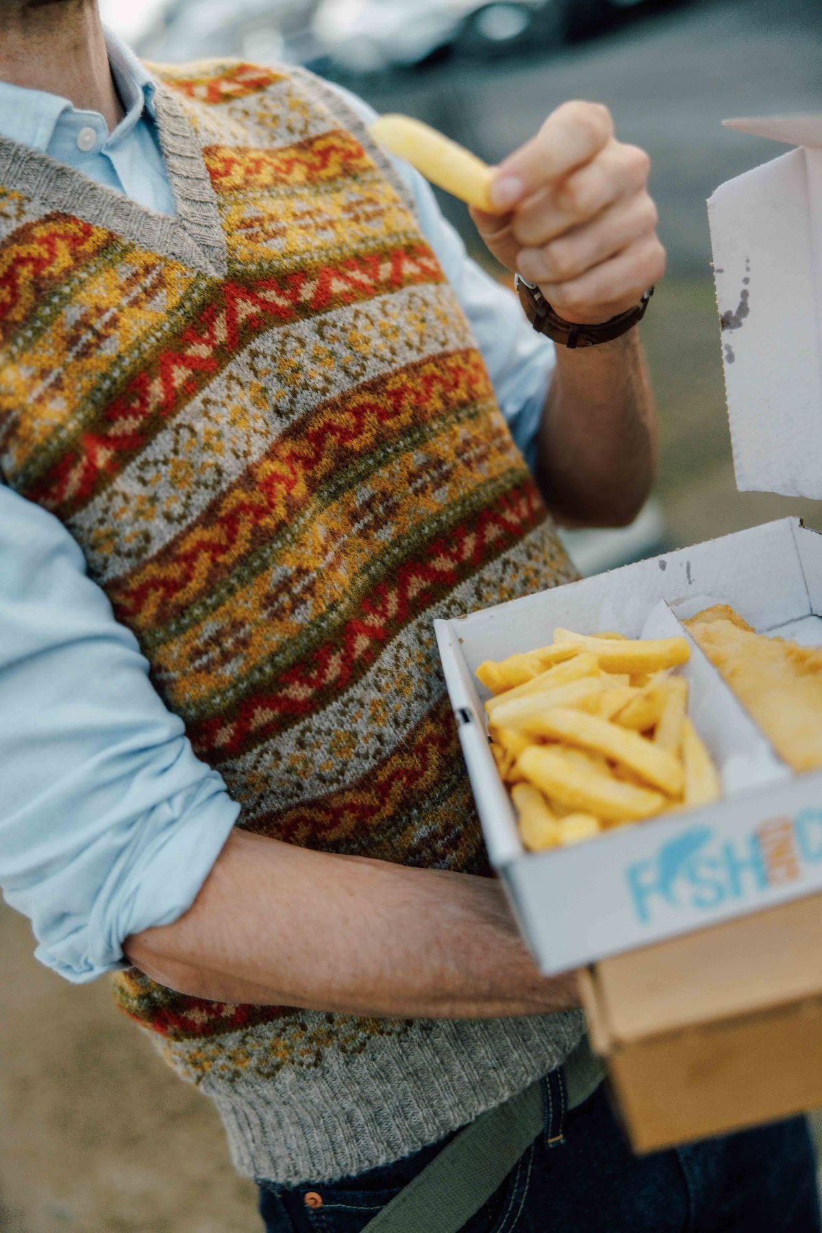 A person wearing a patterned sweater vest holds a box of fish and chips outdoors, picking up a fry with one hand. The background is blurred, focusing on the food and the persons torso.