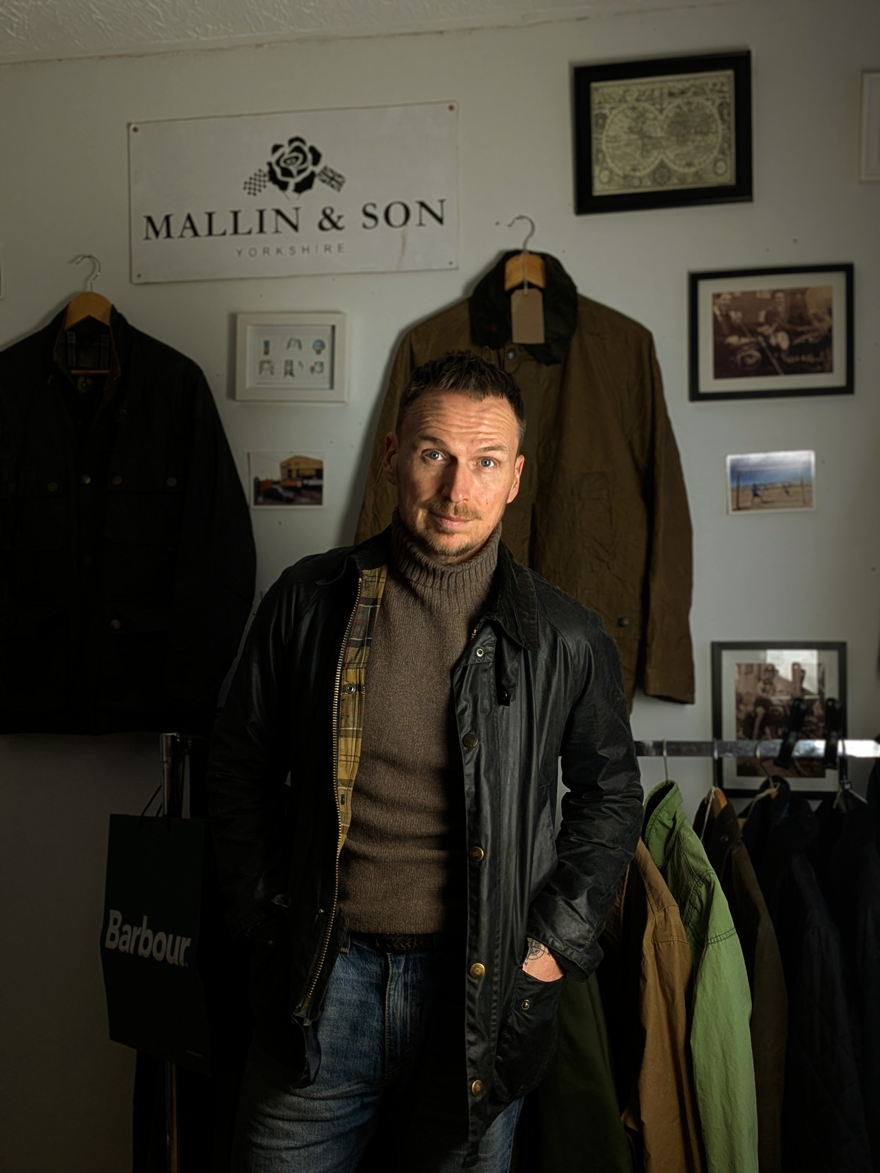 A man in a brown turtleneck and black jacket stands in a shop with coats hanging on racks and framed photos on the wall. A sign reads “Mallin & Son Yorkshire.”.