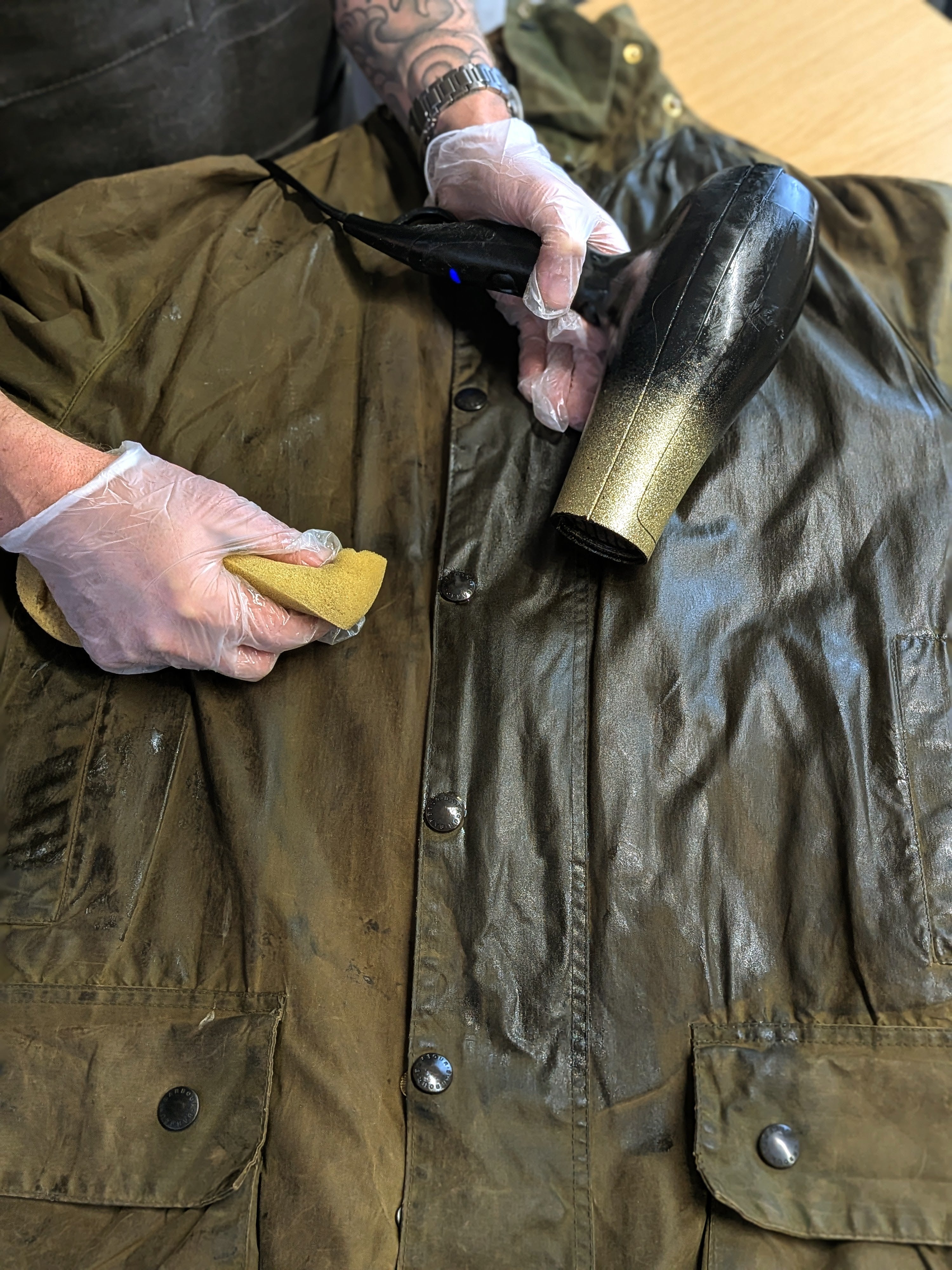 A person wearing gloves uses a sponge and a hairdryer to apply wax to a brown waxed jacket, restoring its finish. The jacket is laid flat on a table, and the persons tattooed arm is visible.