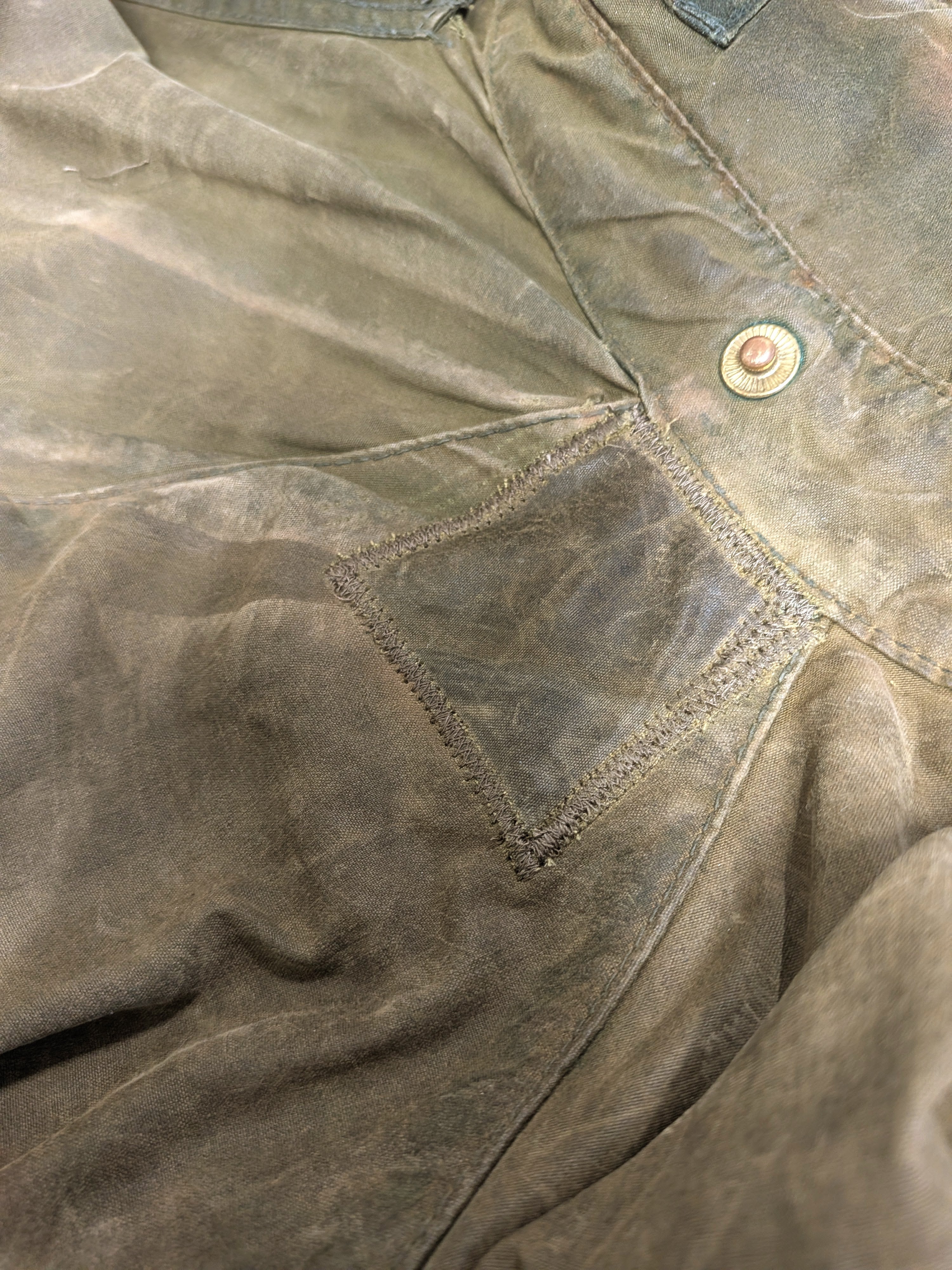A close-up of faded, brownish-green pants with a square leather patch stitched over a worn area near the pocket and a visible button on the waistband.