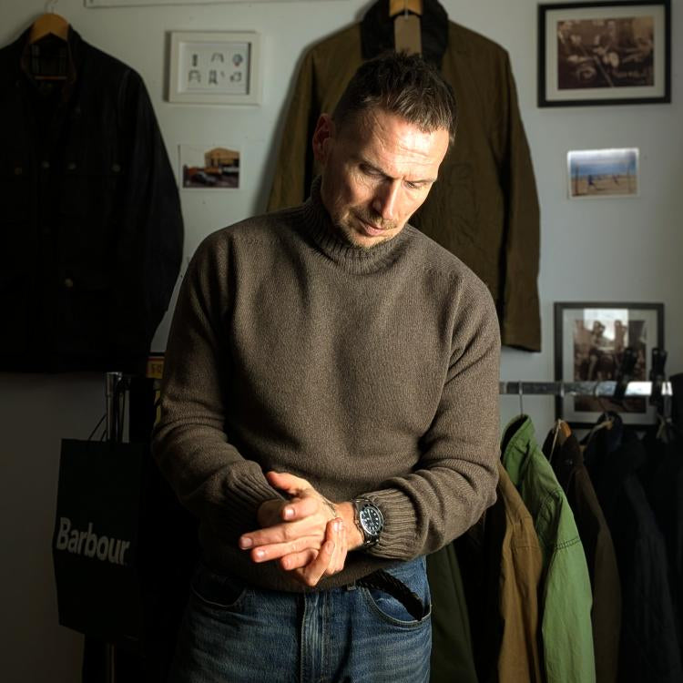 A man wearing a brown sweater and jeans stands indoors, looking down and rubbing his hands. Coats and jackets hang behind him, and framed photos are displayed on the wall. The lighting highlights his face and upper body.