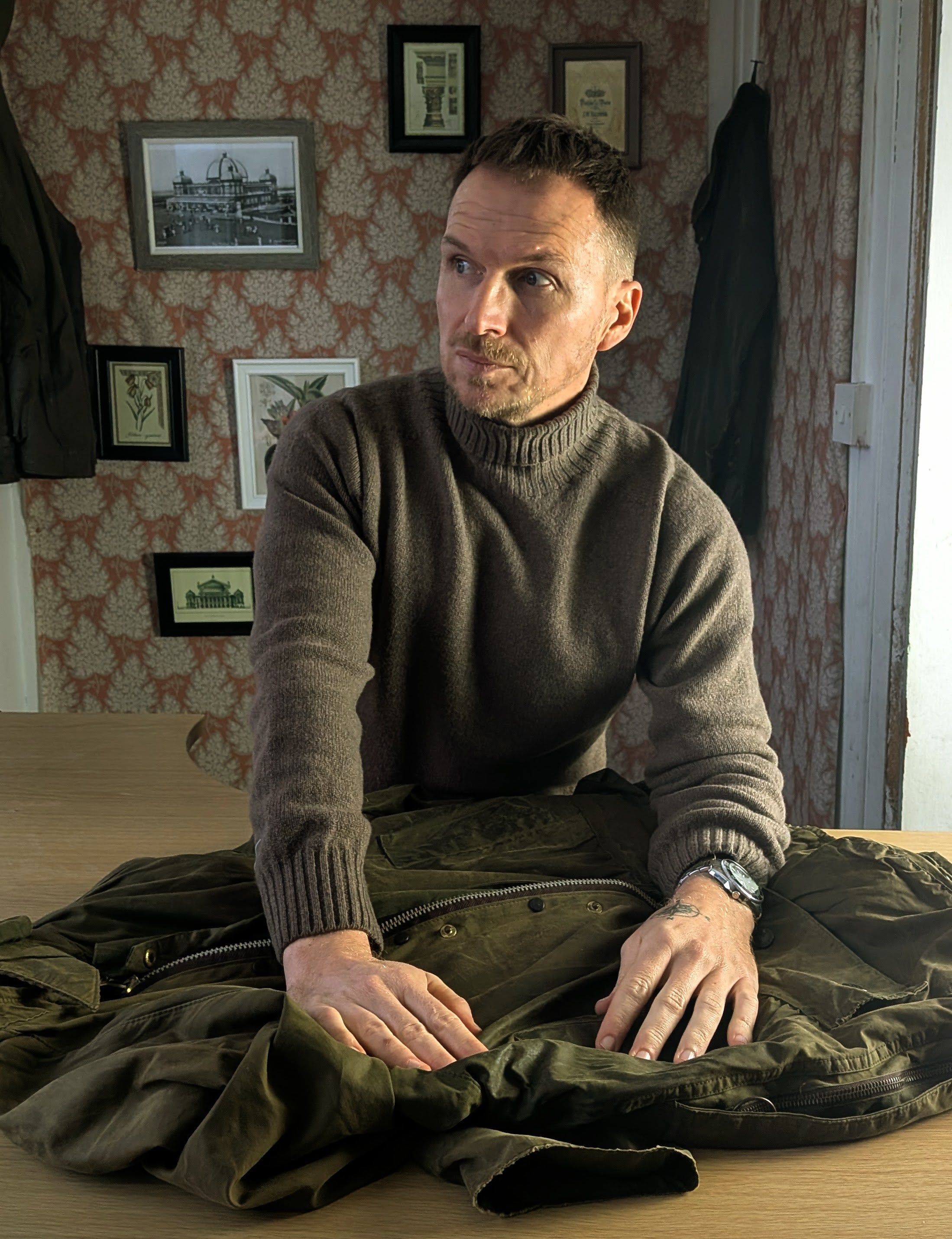 A man in a brown turtleneck sweater stands at a table with a green jacket laid out in front of him. He looks to the side, with framed pictures and patterned wallpaper in the background.