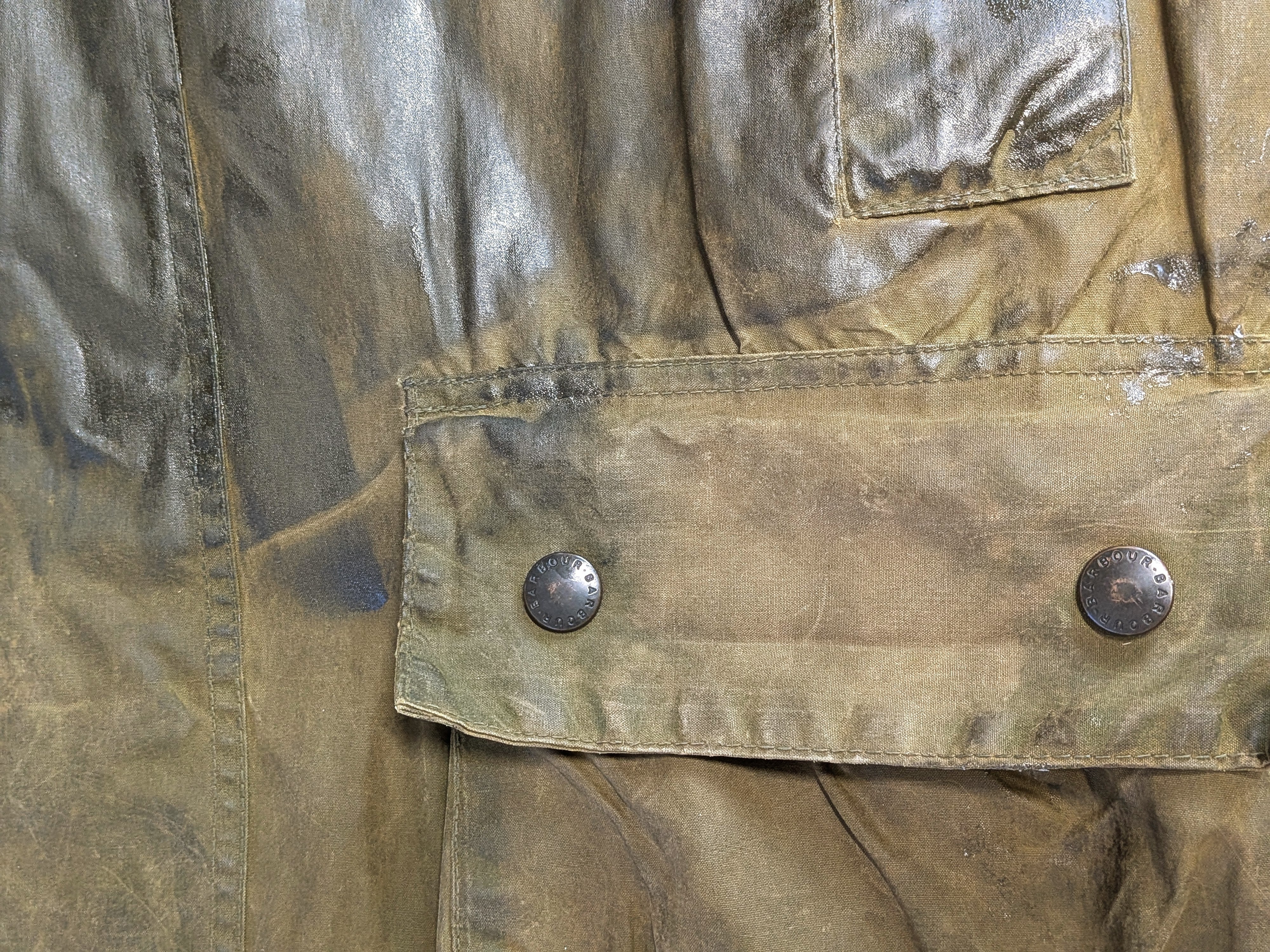 Close-up of a weathered, olive-green fabric jacket showing a flap pocket with two metal snaps and visible seams. The fabric appears worn with slight discoloration and creases.