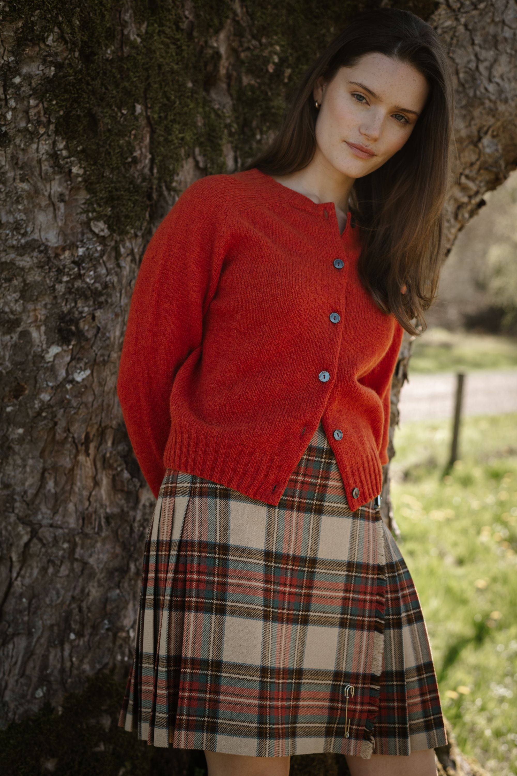 A woman stands outdoors leaning against a tree, wearing a red buttoned cardigan and a pleated plaid skirt. Sunlight filters through the background, highlighting green grass and creating a calm, natural atmosphere.