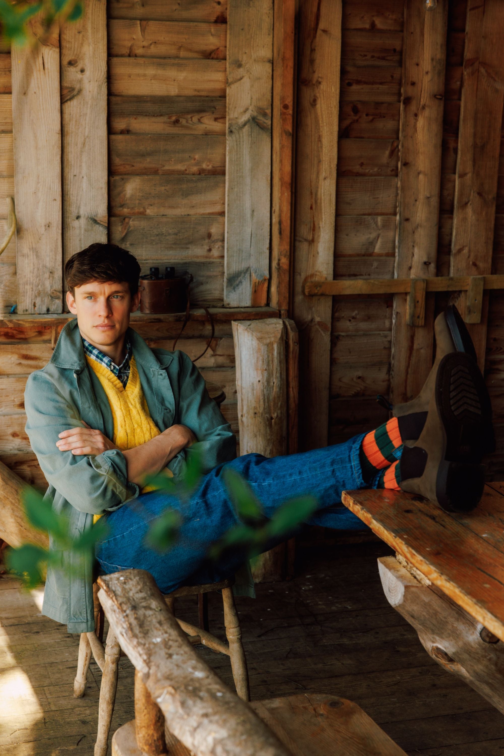 A man in colorful socks, blue jeans, a yellow sweater, and a light jacket sits with his legs up on a wooden table in a rustic wooden cabin, arms crossed and looking at the camera.