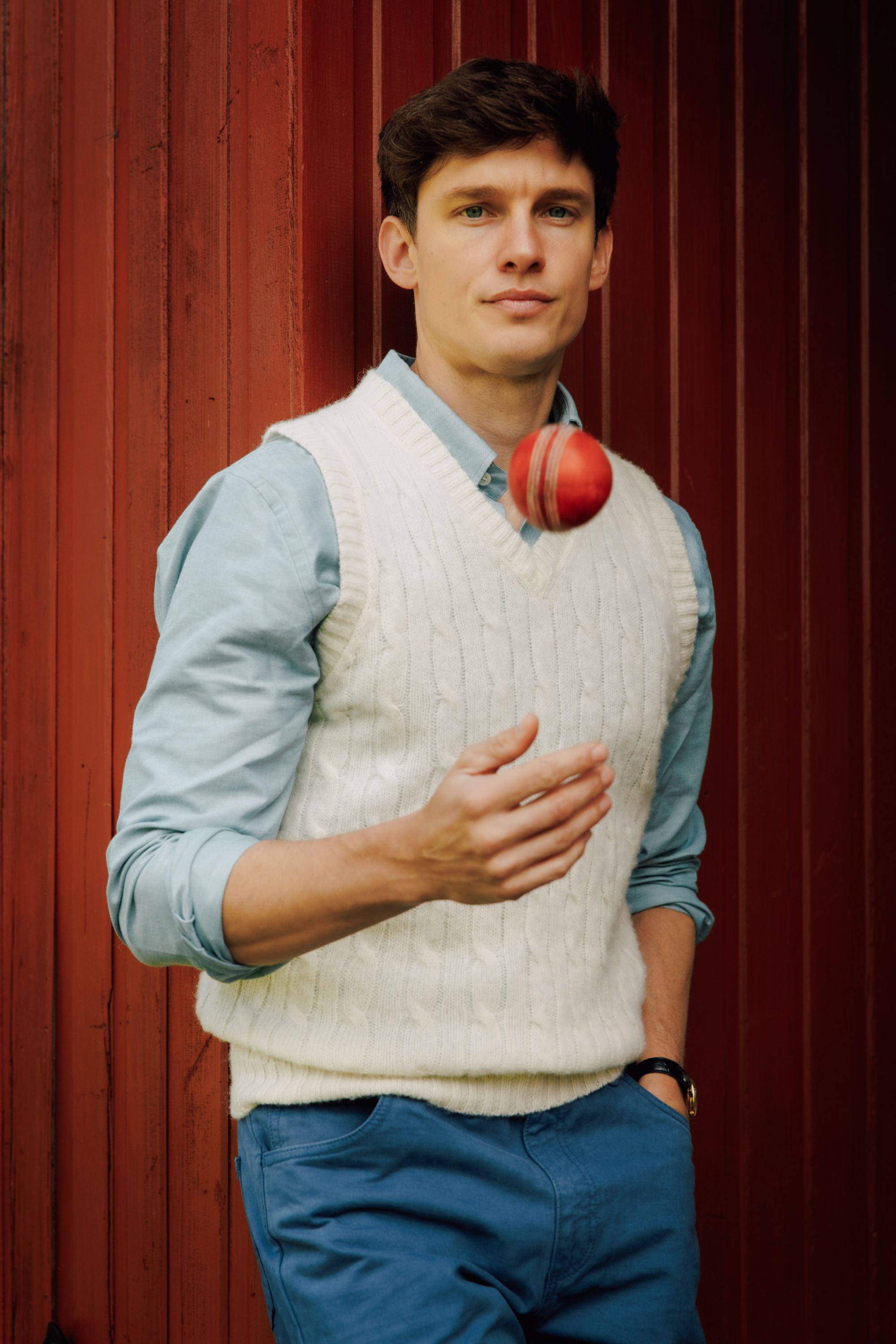A young man in a light blue shirt and white sweater vest stands against a red wall, tossing a red cricket ball in the air with one hand. He is looking at the camera and smiling slightly.