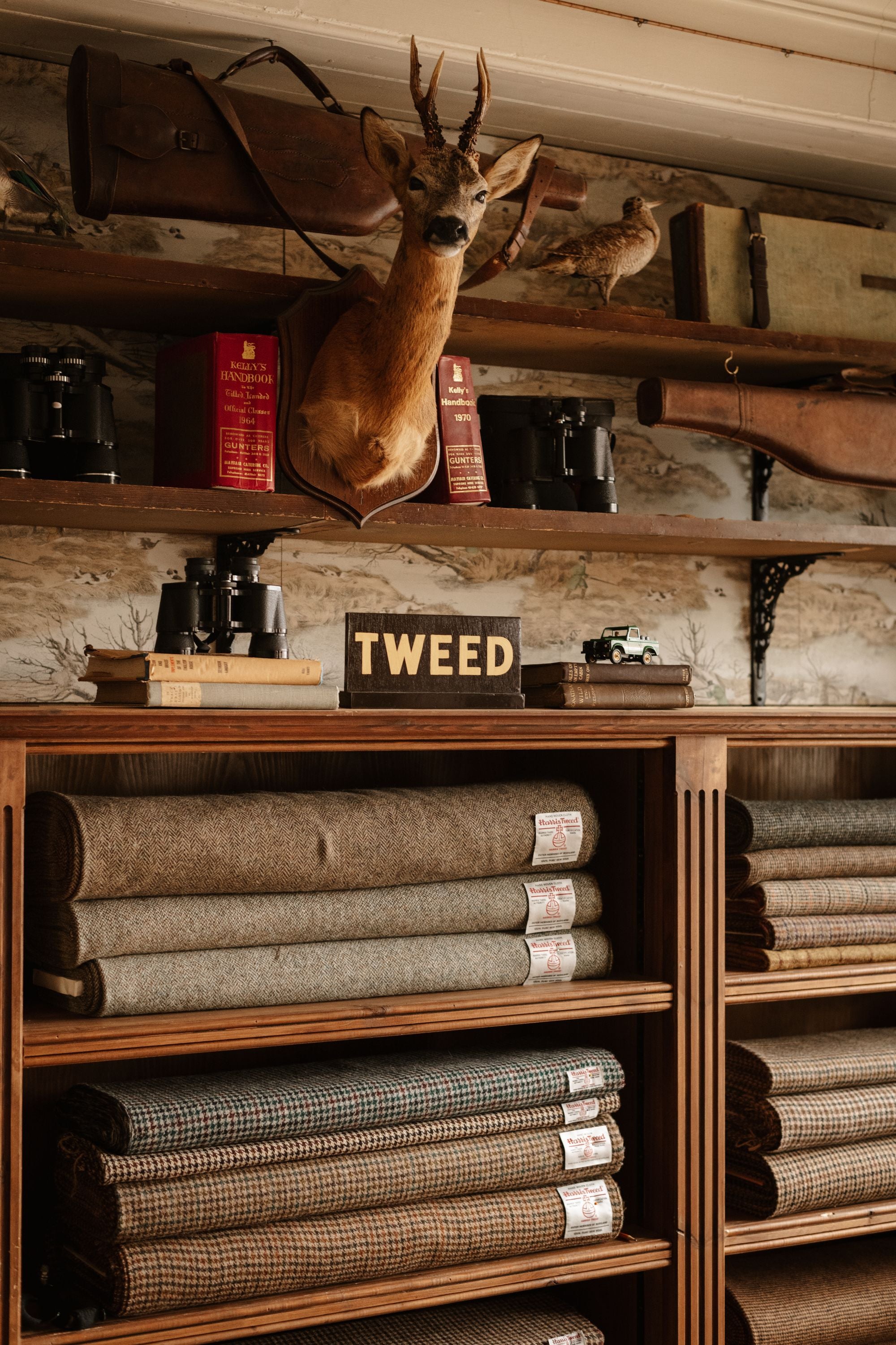 Shelves display neatly folded tweed fabric rolls, vintage books, binoculars, and a wooden sign labeled “TWEED.” Above, a mounted deer head and rustic decor add to the shop’s classic, cozy atmosphere.