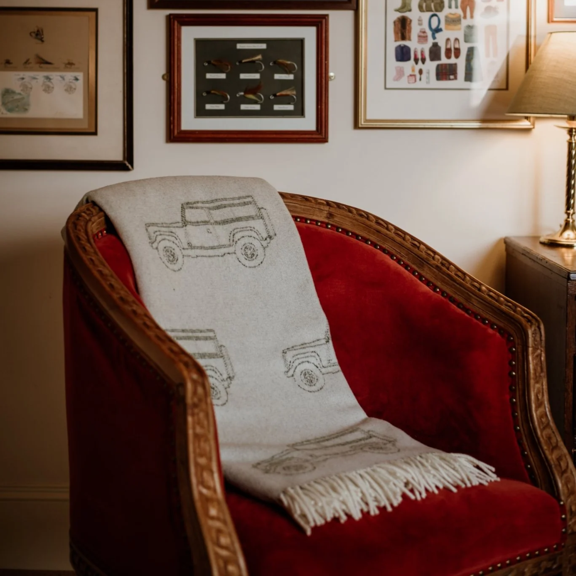 A wooden armchair with red upholstery is draped with a light grey fringed blanket featuring vehicle outlines. The chair is next to a lamp and in front of a wall decorated with various framed pictures.