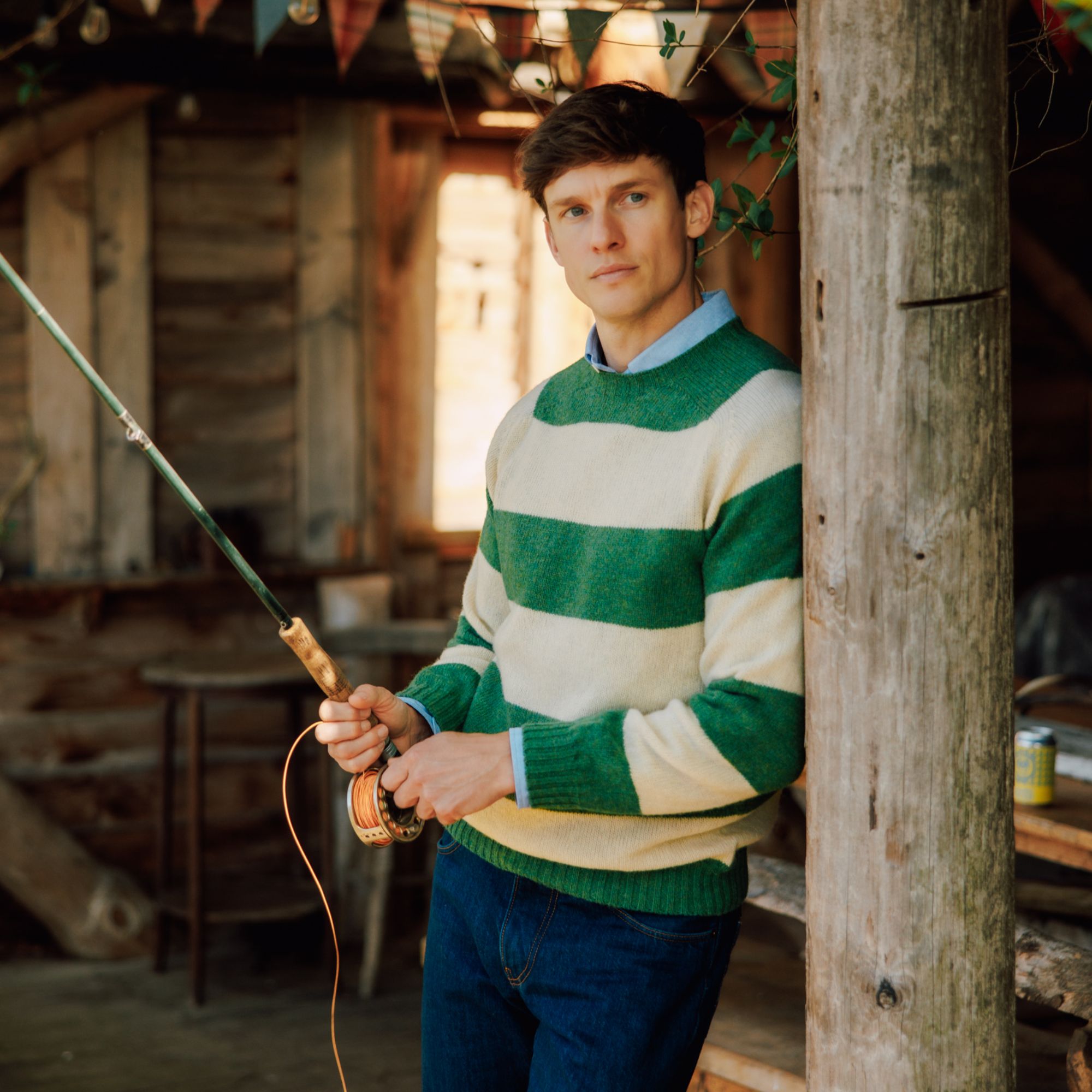 A person wearing a green and white striped sweater and jeans stands indoors, leaning against a wooden post, holding a fishing rod. The background is rustic with wooden walls and string lights.