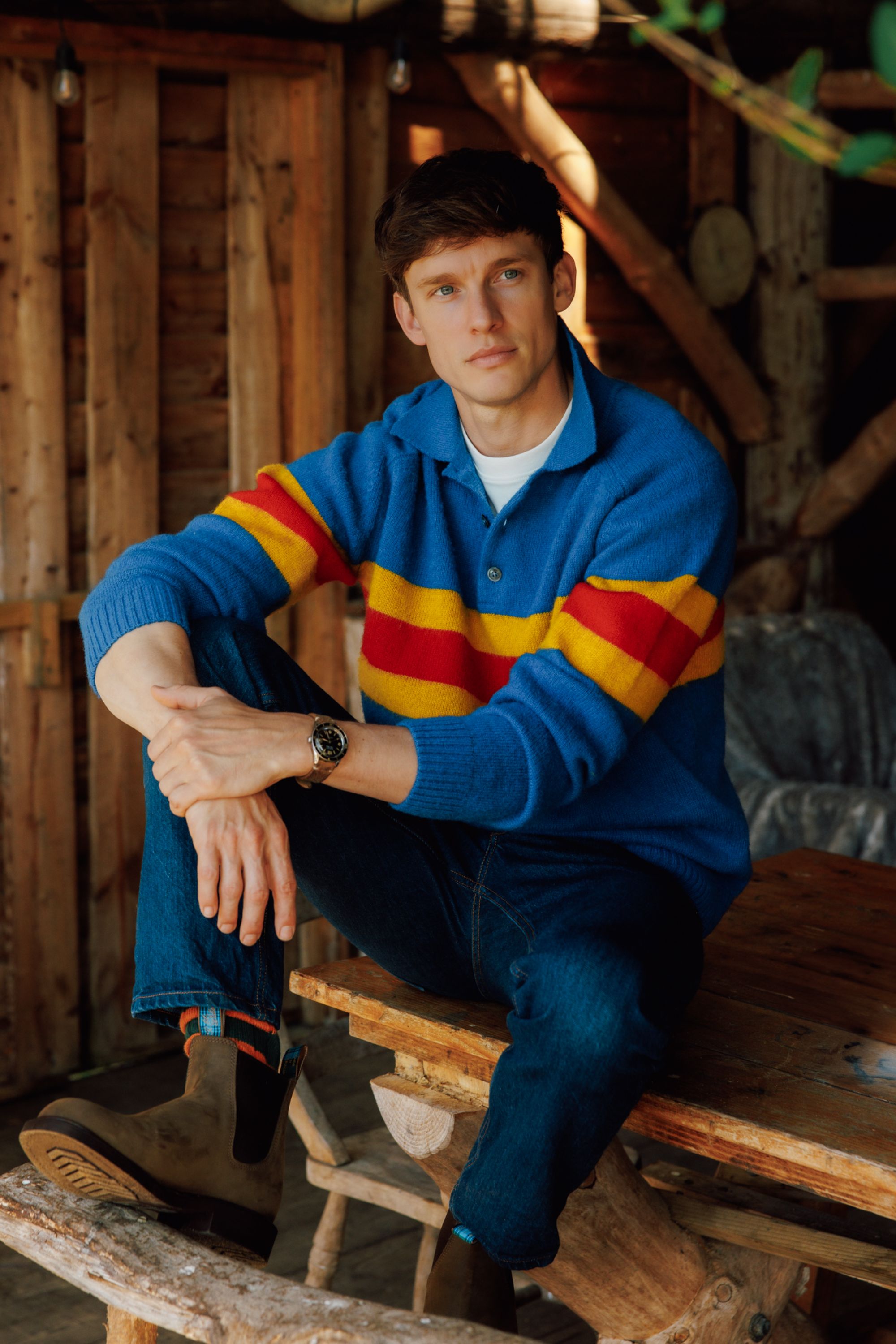 A young man in a blue sweater with red and yellow stripes sits on a rustic wooden table in a cozy, wood-paneled cabin. He has short brown hair and wears jeans and brown boots, looking thoughtfully at the camera.