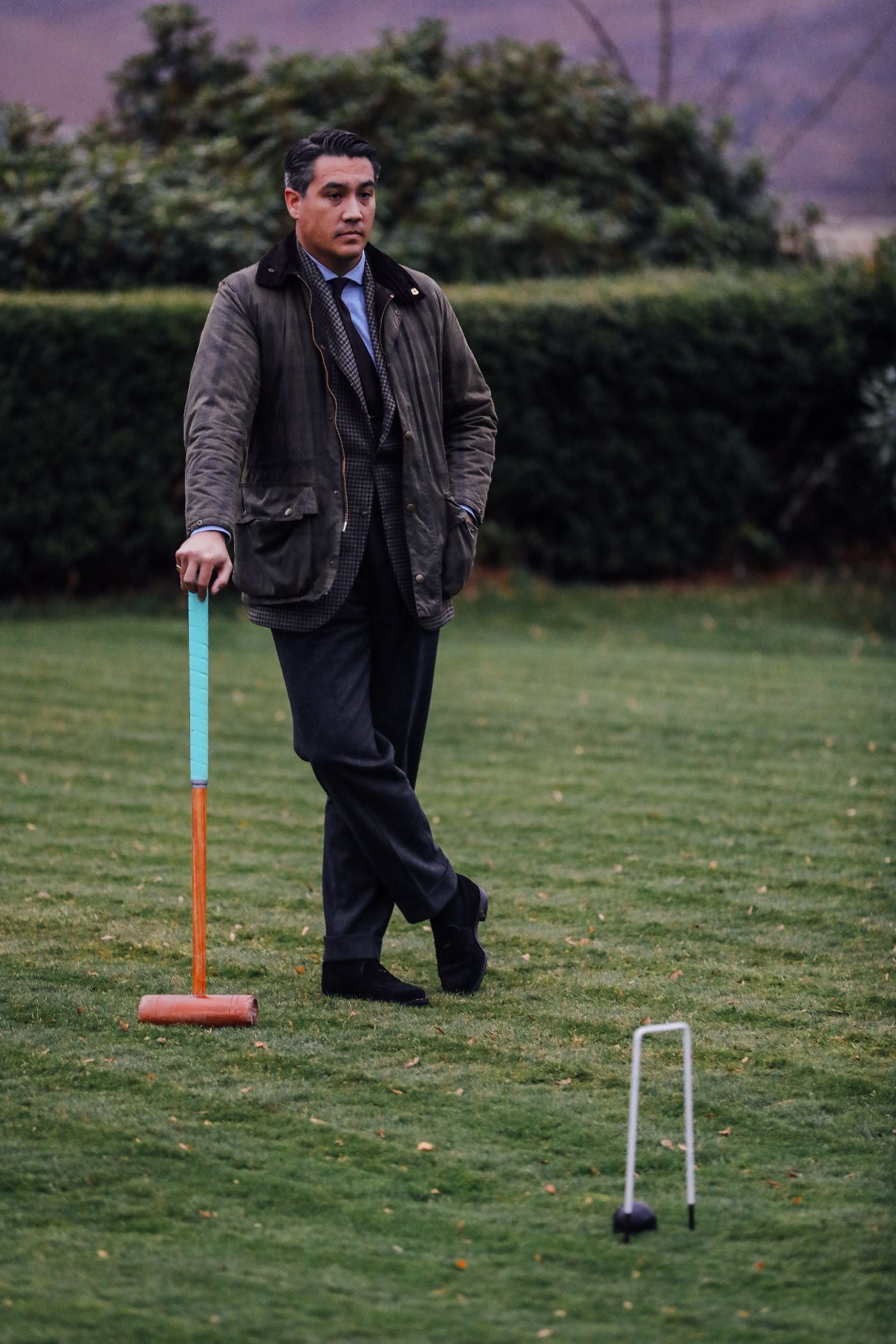 A man in a jacket and tie stands on a grassy lawn, leaning on a croquet mallet. A croquet wicket and ball are set up in front of him, with green hedges in the background.