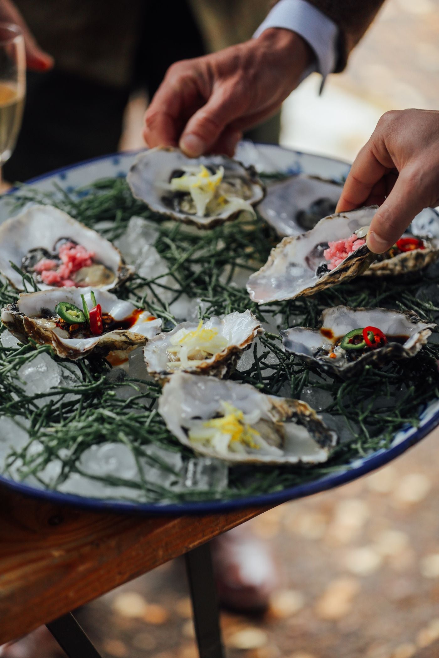 A platter of oysters on a bed of ice and seaweed, garnished with lemon zest, red chilies, and other toppings. Two hands are reaching in to take oysters. A glass of wine is partially visible in the background.