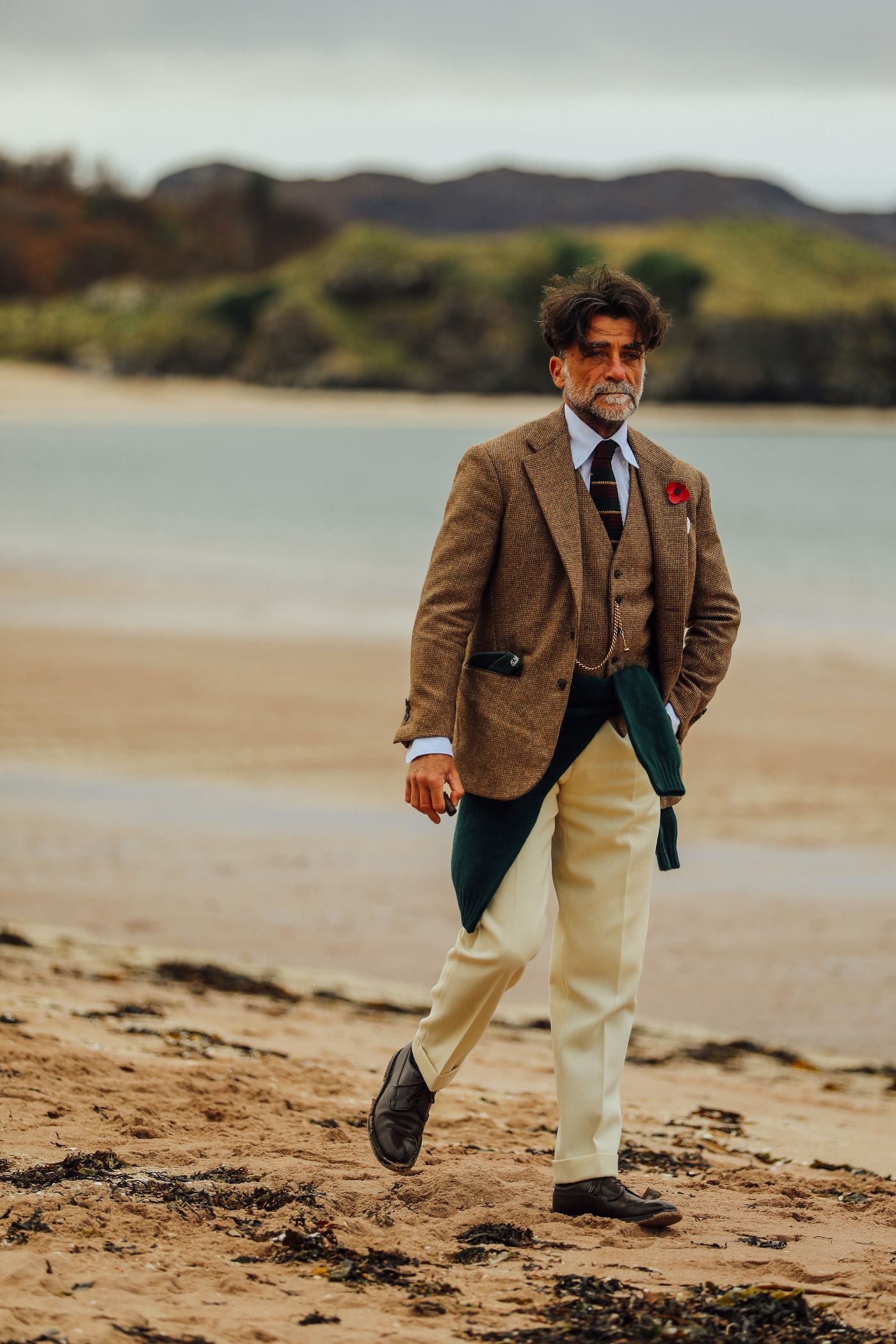A man wearing a brown blazer, waistcoat, white shirt, striped tie, and cream trousers walks on a sandy beach with grassy dunes and hills in the background.