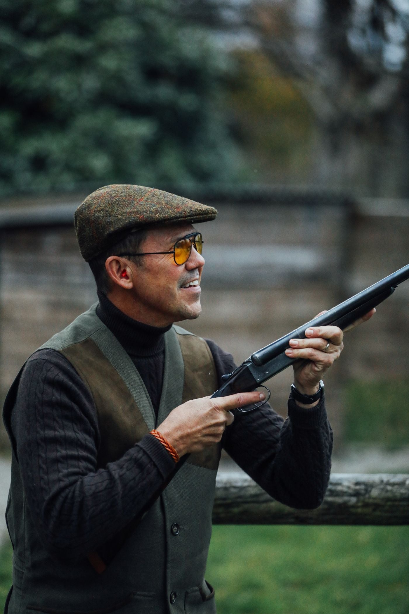 A man wearing a brown flat cap, yellow-tinted glasses, and a dark sweater aims a shotgun outdoors, smiling. There is greenery and a wooden fence in the background.