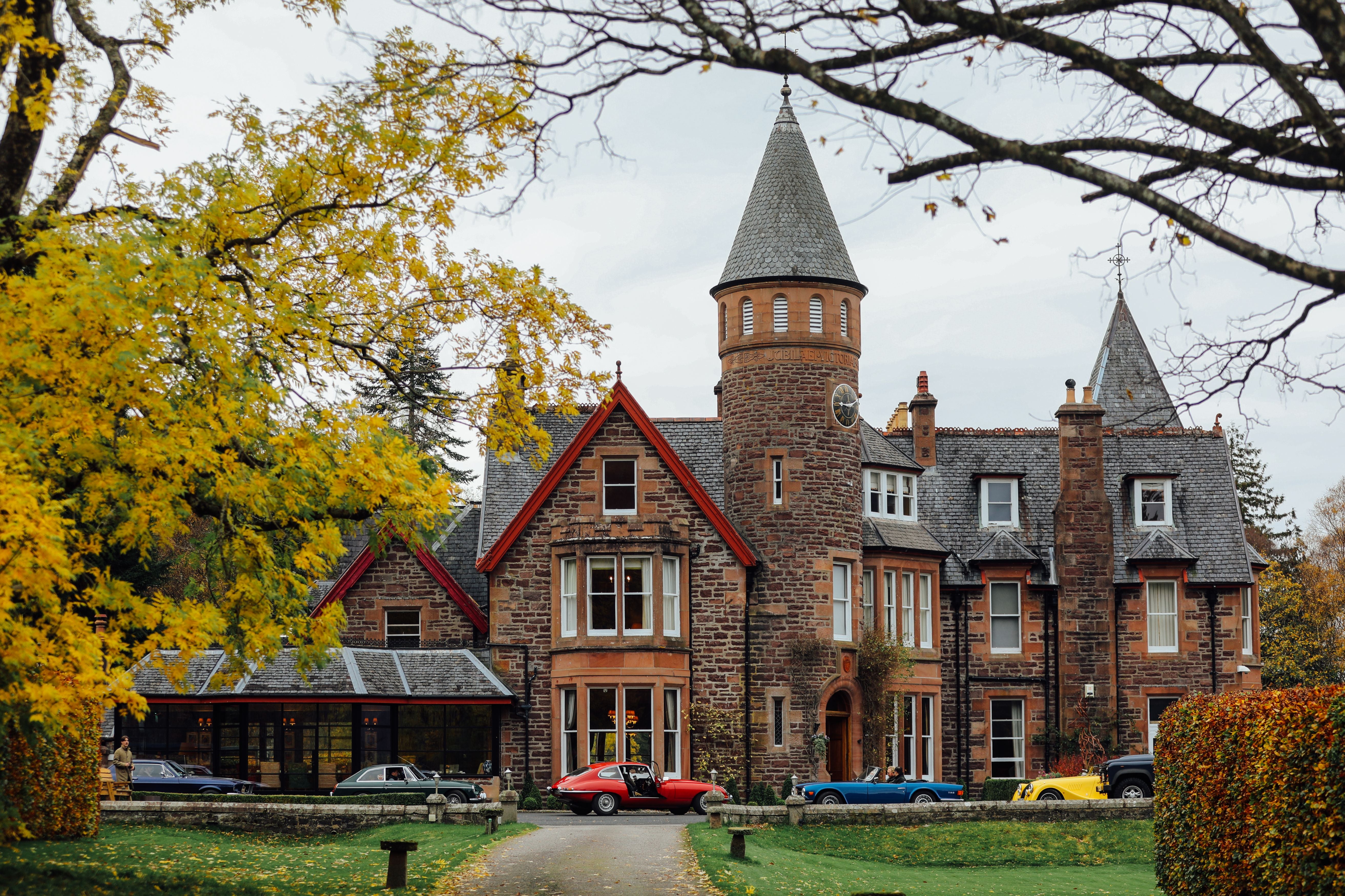 A large, historic stone mansion with turrets and slate roofs, surrounded by autumn trees. Several classic cars, including a yellow and a red one, are parked on the driveway in front of the house.