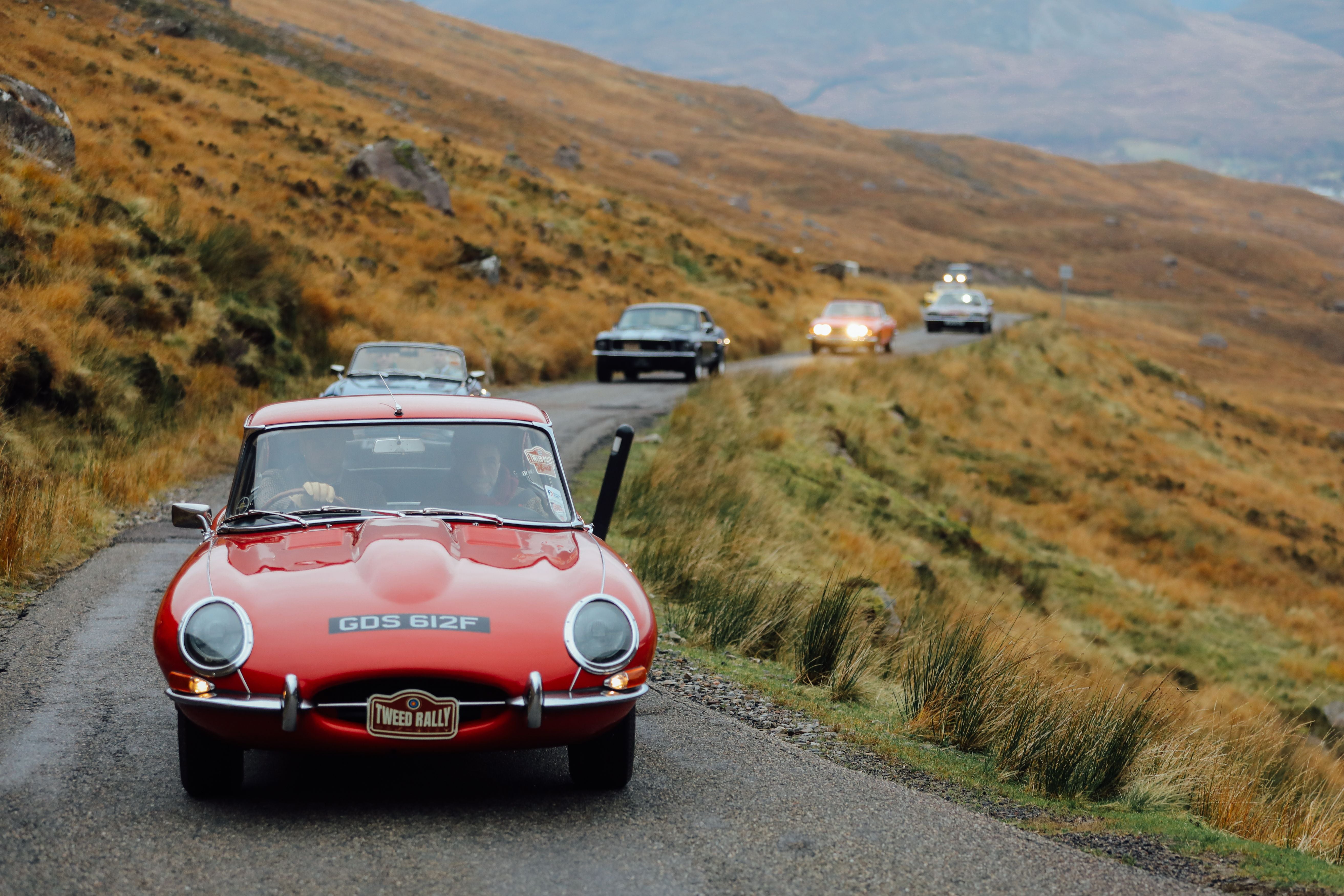 A convoy of classic cars drives along a winding, narrow road through a hilly, grassy landscape. The lead car is a red vintage Jaguar with the license plate GDS 612F.