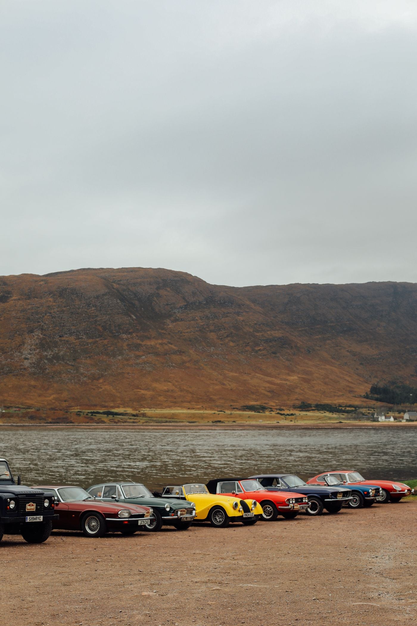 A row of vintage cars in various colors is parked on a gravel area beside a lake, with hills covered in brown grass in the background under a cloudy sky.