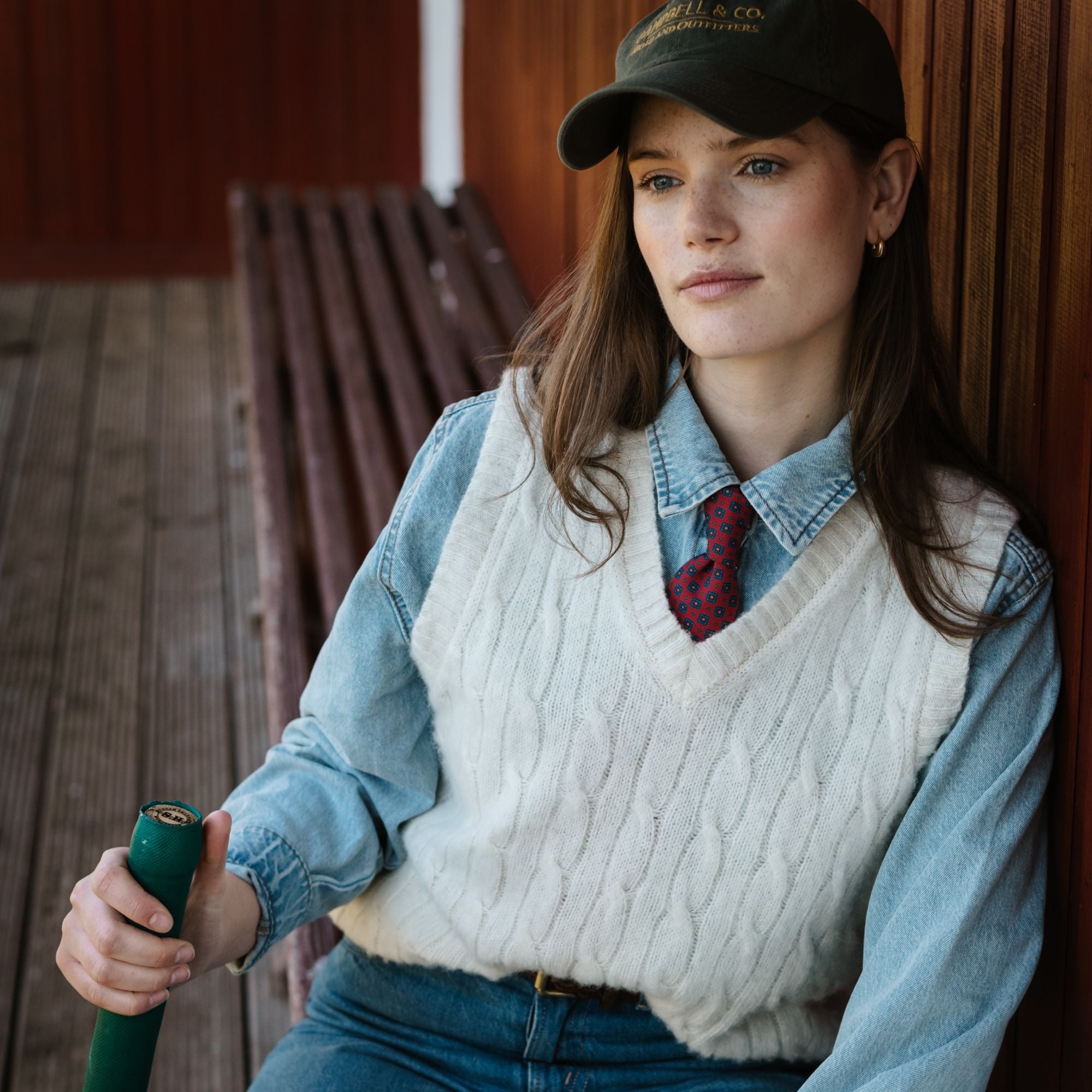 A woman in a cap, denim shirt, patterned tie, and white cable-knit vest sits on a wooden bench, holding a green-handled object, looking thoughtful. The background features wooden slats and floorboards.