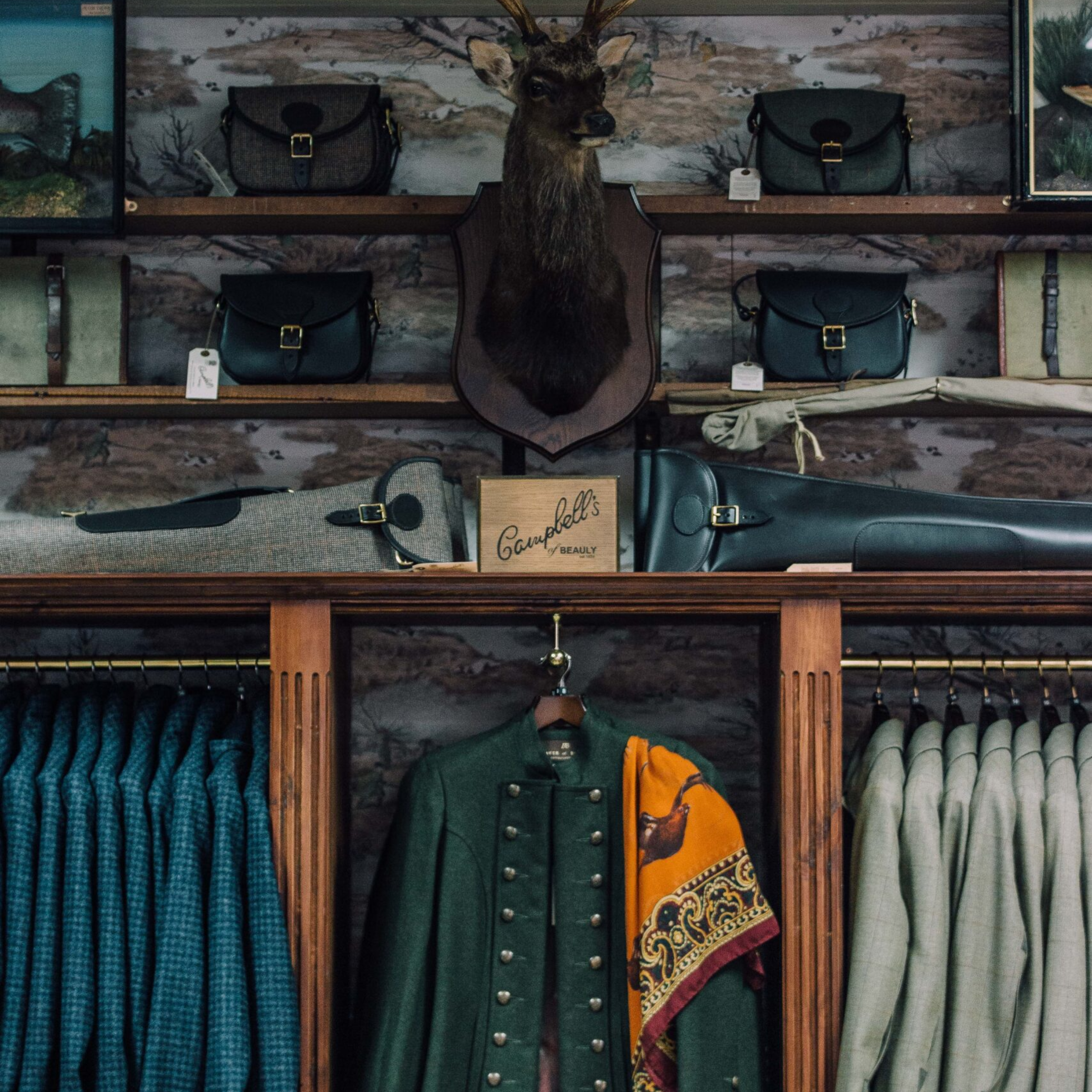 A clothing store display featuring neatly hung blue and grey jackets, a green coat with a colorful scarf, shelves of handbags, cases, and a mounted deer head above the display.