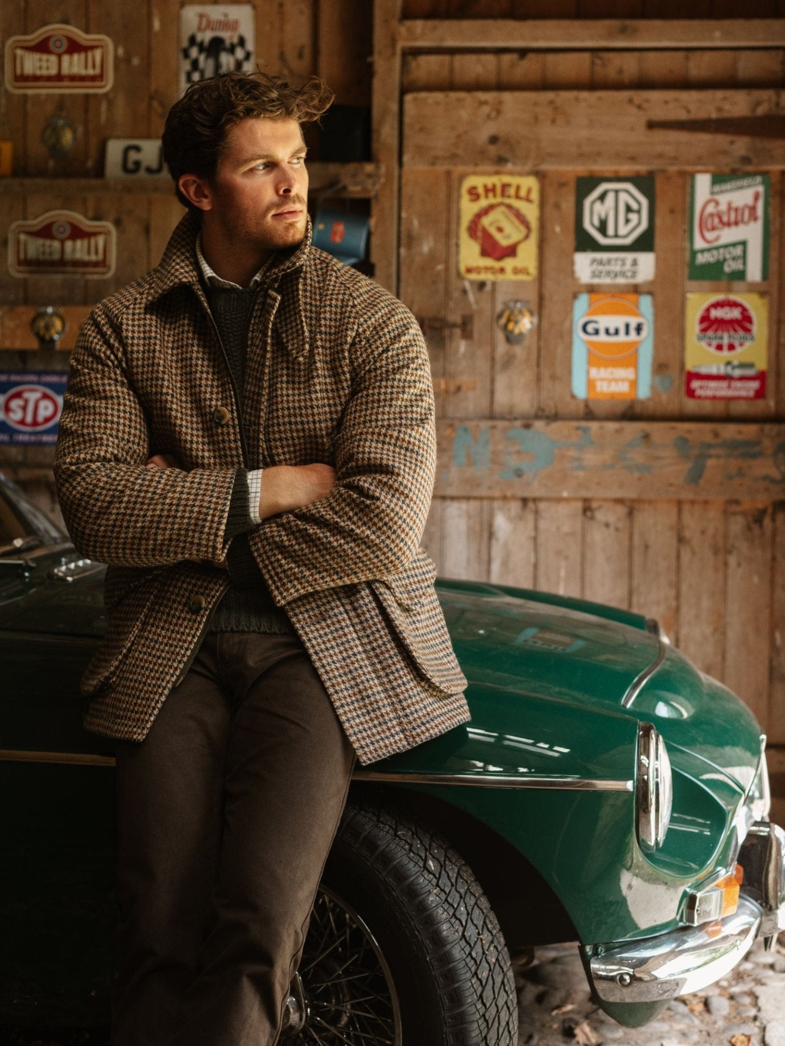A man in the Cannich Coat by Campbell's of Beauly leans against a classic green car in a rustic garage filled with vintage automotive signs, looking to the side with his arms crossed.