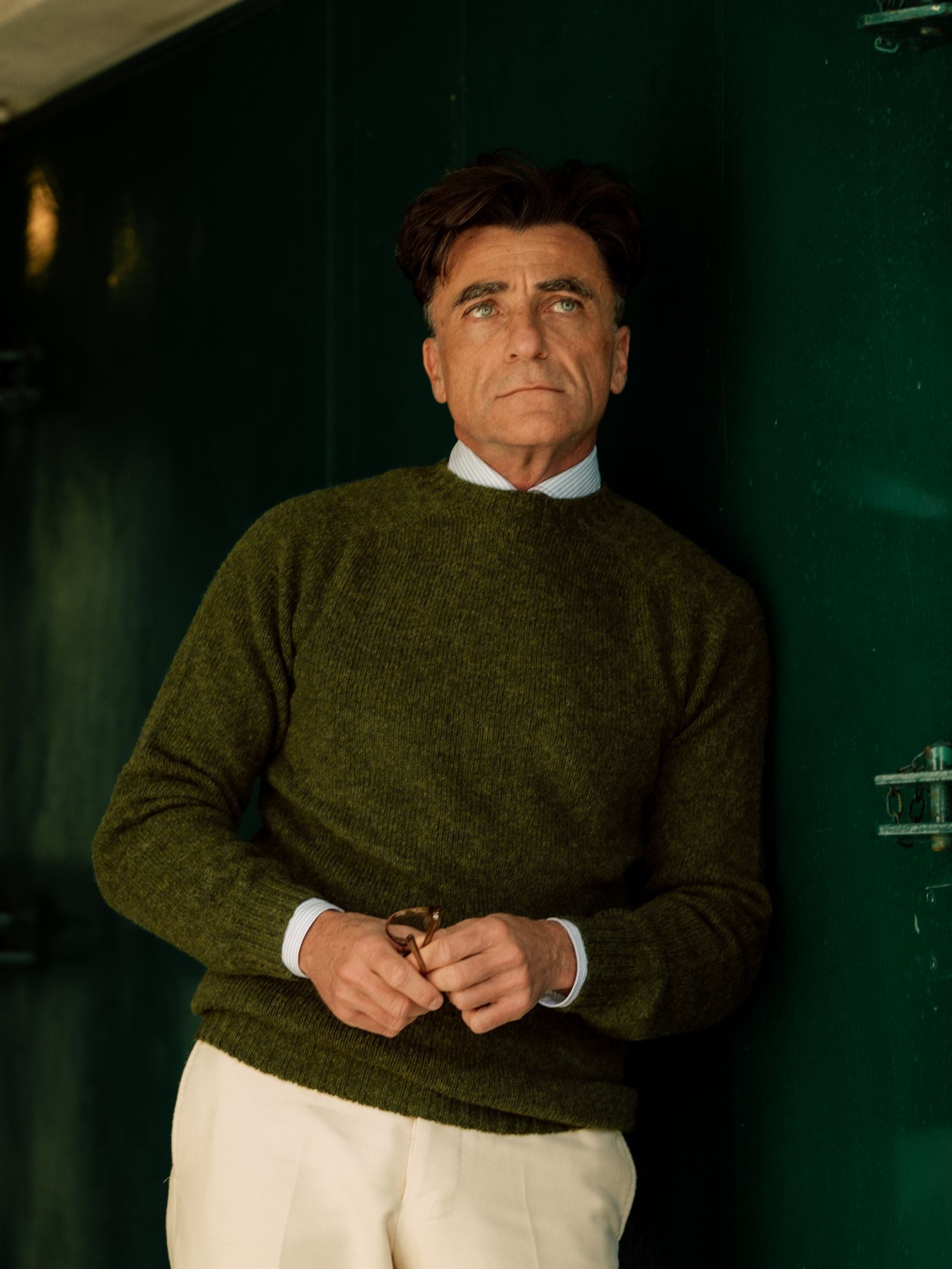 A man with dark hair wears the Campbell's of Beauly Shetland Jumper in green over a collared shirt and cream pants, standing against a dark green wall, holding eyeglasses and gazing slightly upward.