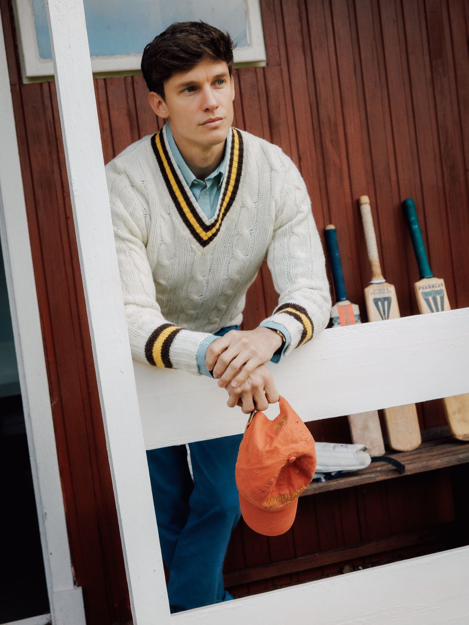 A young man in a Campbell's of Beauly Shetland Cricket Sweater leans on a white fence, holding an orange cap, with cricket bats behind him against a red wooden wall and bench. He looks thoughtful, gazing into the distance.