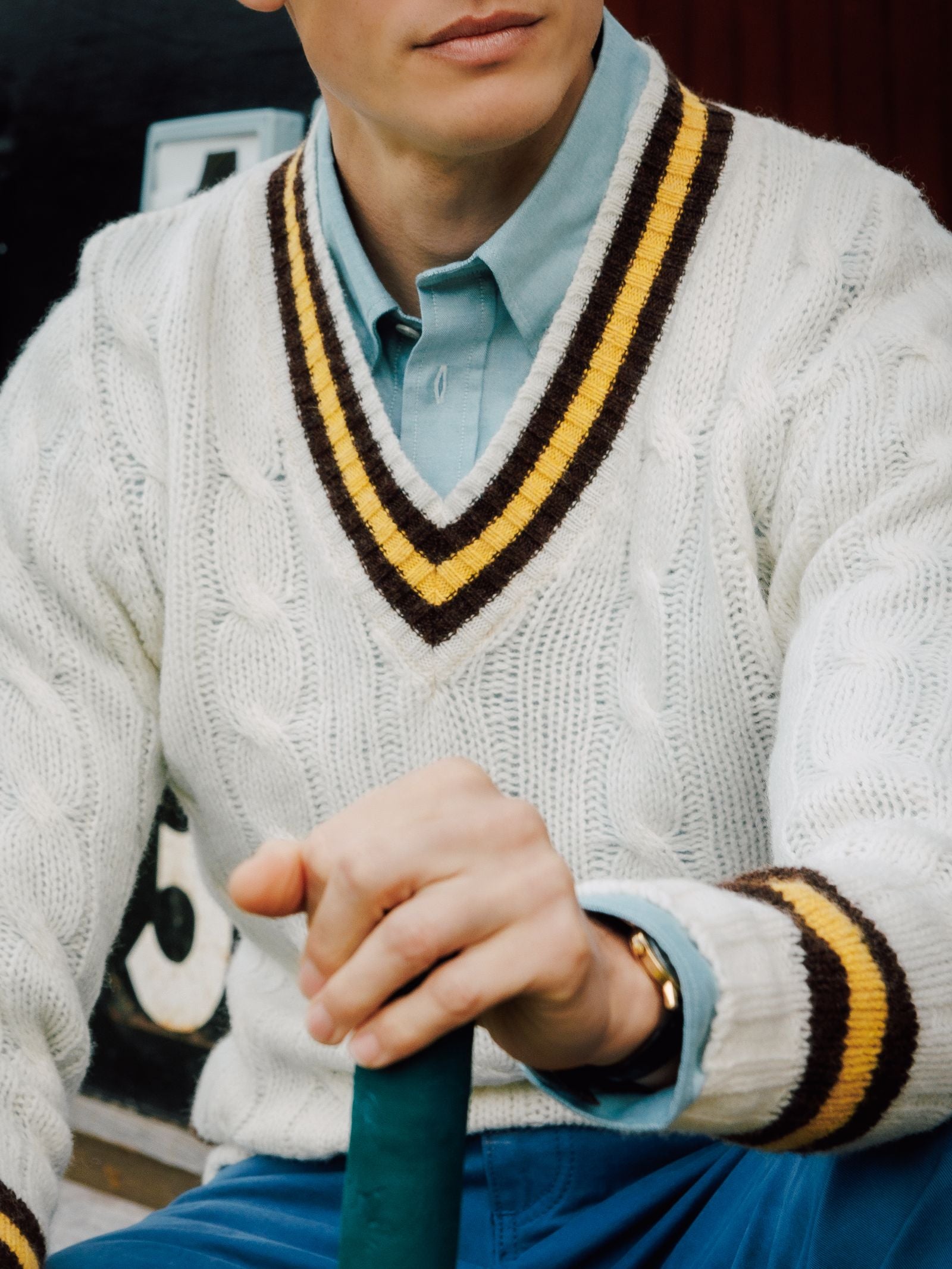 A person wears a Campbell's of Beauly Shetland Cricket Sweater with yellow and brown stripes over a light blue collared shirt, holding a green item, possibly a bat, with part of a scoreboard in the background.