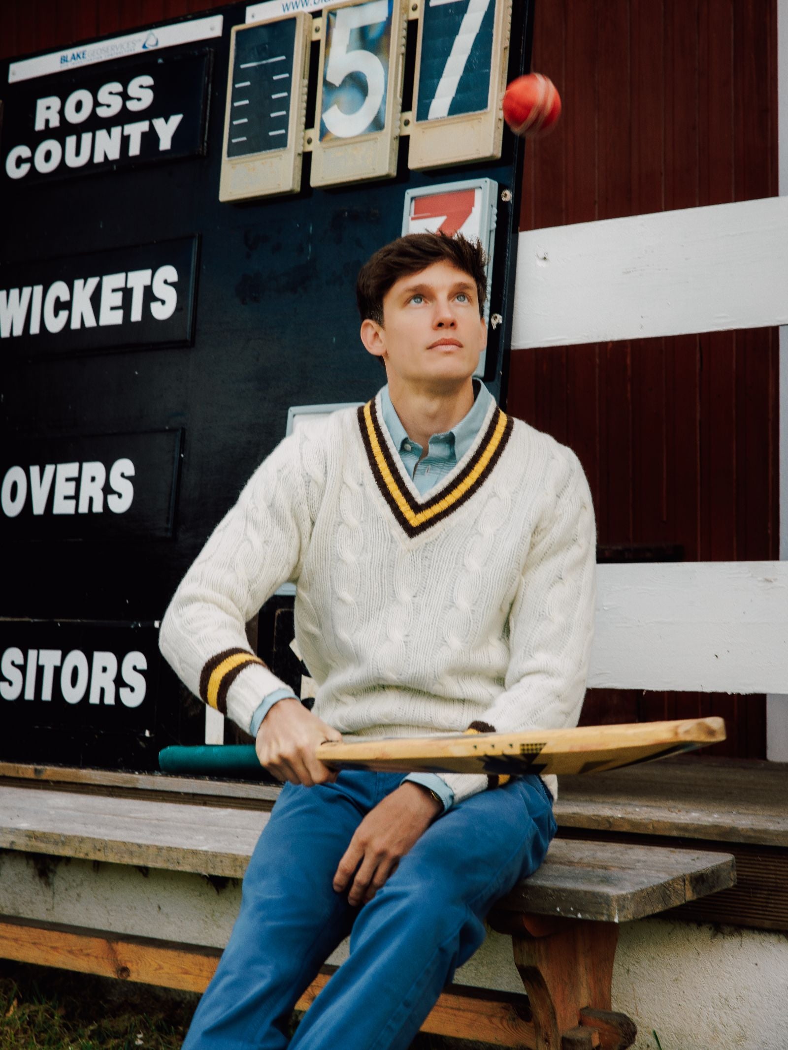 A young man wearing the Campbell's of Beauly Shetland Cricket Sweater and blue pants sits on a bench, holding a cricket bat and tossing a red ball, with a scoreboard visible in the background.
