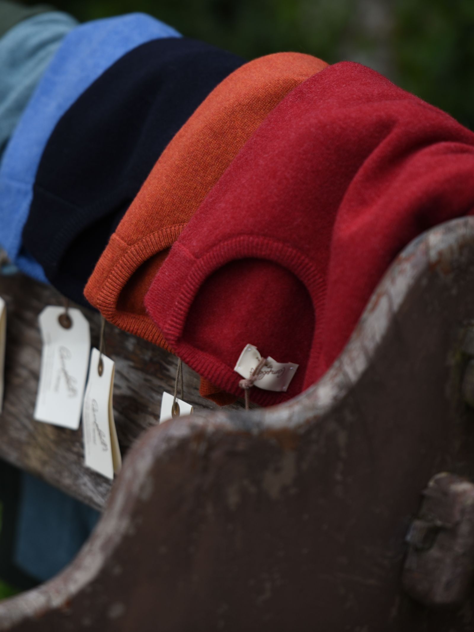 Four folded Campbell’s of Beauly Auld Stock Lambswool Crew Neck Jumpers in blue, black, orange, and red are stacked on a weathered wooden bench, each with a white tag attached for sale or display.