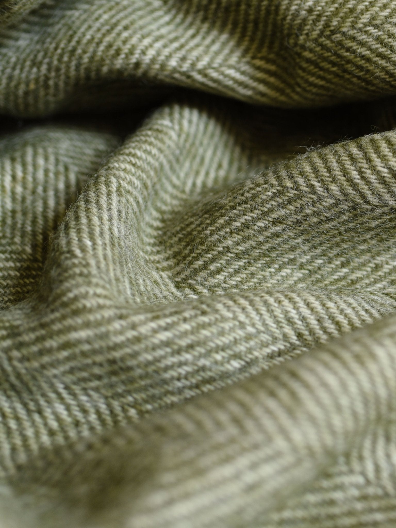 Close-up of the British Wool Green Throw by Campbell's of Beauly, showing its soft, folded fabric in a green and beige herringbone pattern. The Scottish-woven wool texture adds a cozy and warm look.