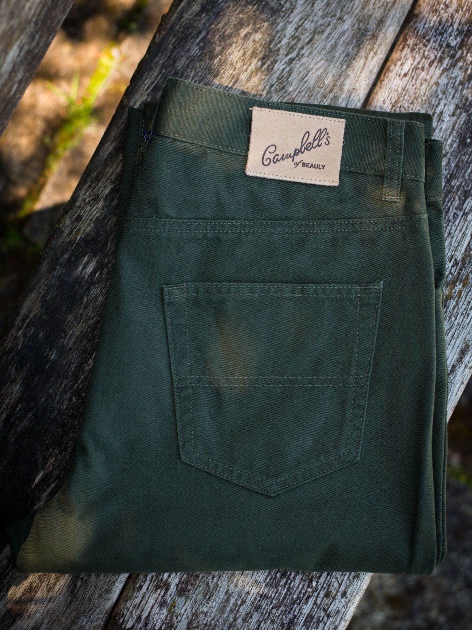 A pair of dark green Colour Cotton Jeans by Campbell's of Beauly, straight leg style, neatly folded on a weathered wooden surface.