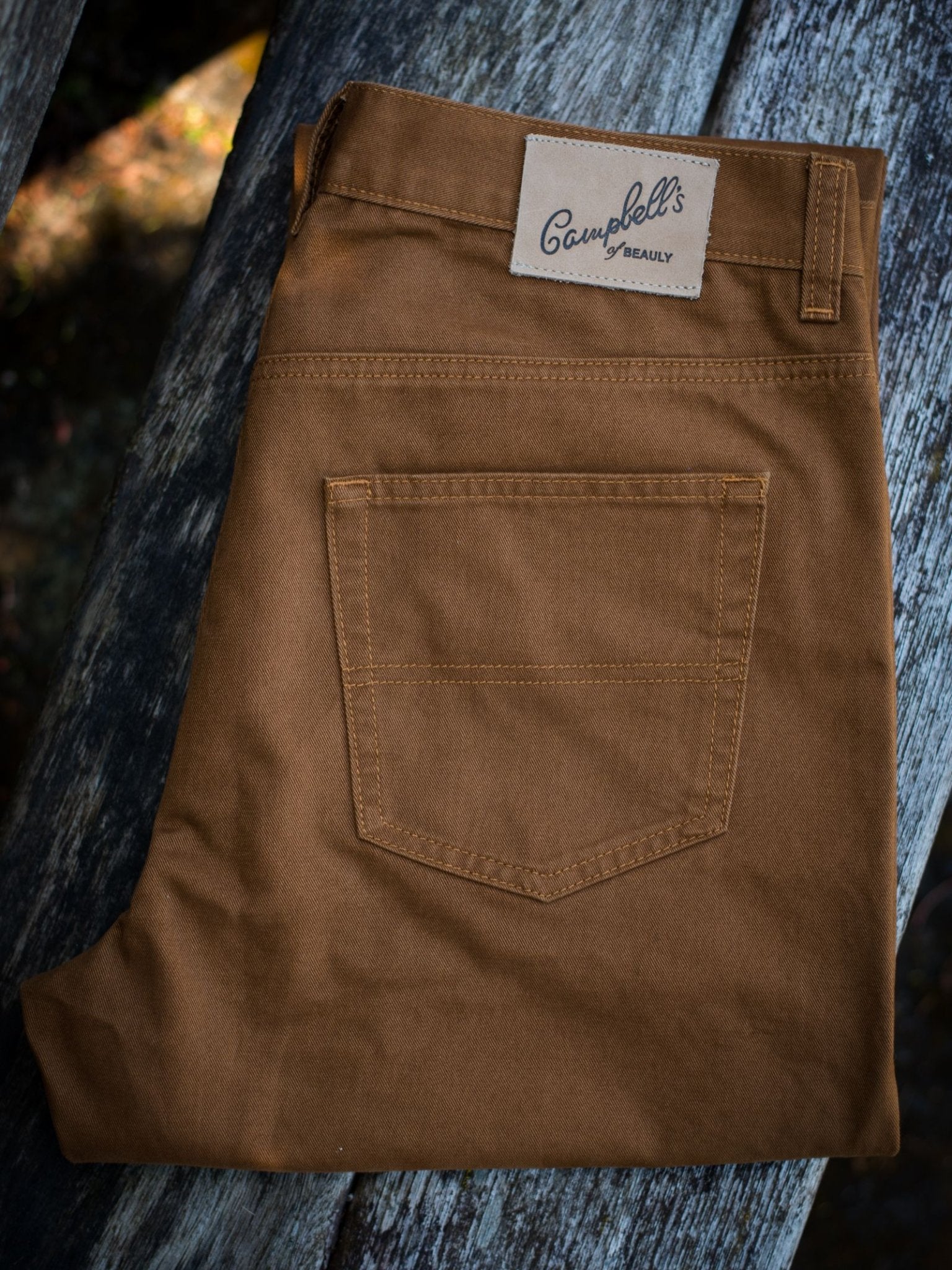 A pair of Colour Cotton Jeans from Campbell's of Beauly, in brown with straight legs, folded neatly to show the back pocket and beige brand label, rests on a weathered wooden surface.
