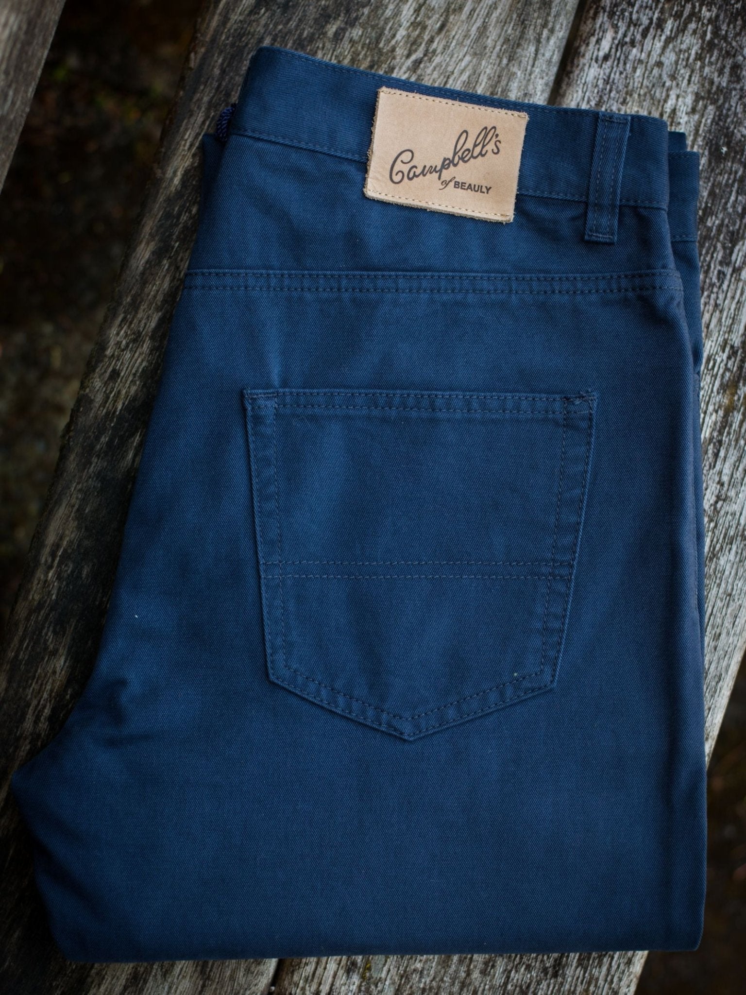 A pair of folded Colour Cotton Jeans by Campbell's of Beauly, featuring a classic five-pocket design and a brown branded label on the waistband, displayed on a weathered wooden surface.