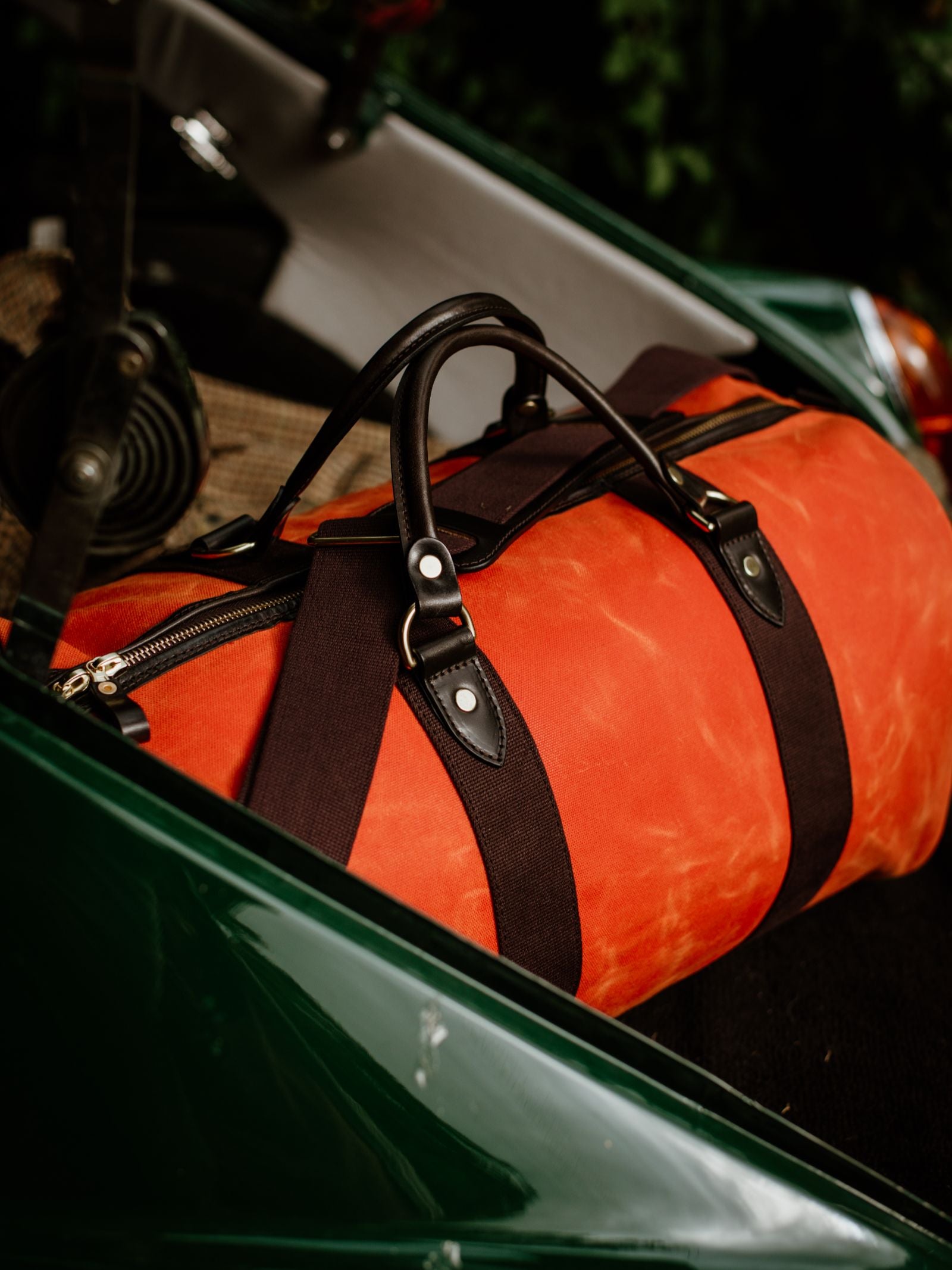 The Campbell's of Beauly Wax Holdall, an orange travel bag with black straps and bridle leather handles, rests on the seat of a green vehicle partially visible in the background.