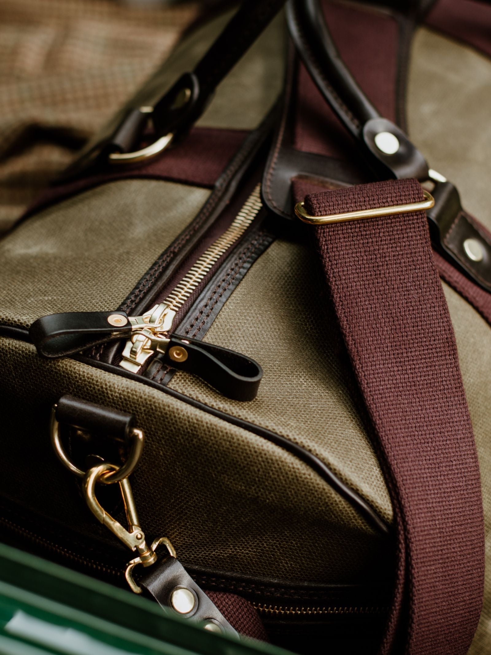 Close-up of Campbell's of Beauly Wax Holdall, a green waxed canvas duffel with brown leather accents, bridle leather handles, gold zipper, maroon shoulder strap with brass hardware, resting on a plaid fabric surface.