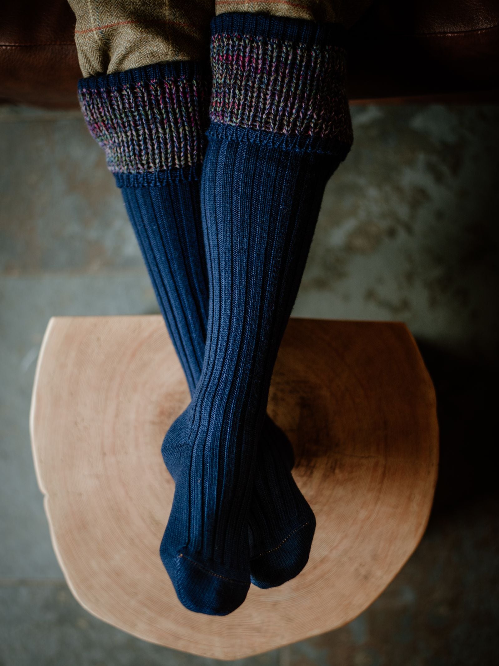 Person wearing Campbell's of Beauly Auld Stock Merino Katrine Shooting Socks—navy blue, ribbed, with multicolored cuffs, handmade in Scotland—sits cross-legged on a wooden stool, viewed from above.