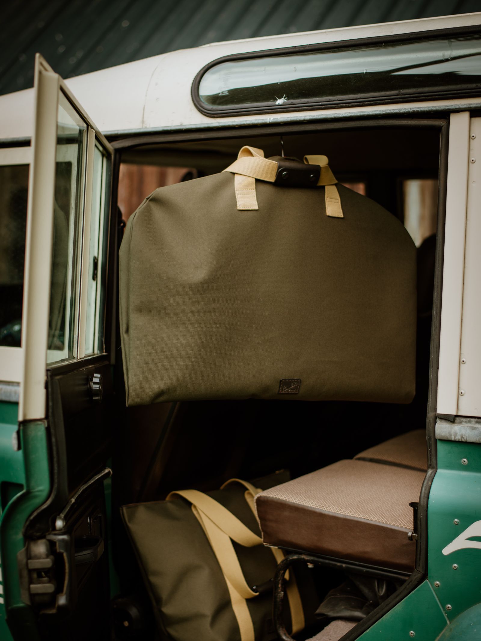 An olive green Campbell's of Beauly Utility Suit Bag, crafted from heavy-duty canvas with beige handles, hangs on a vintage vehicle’s door for stylish garment protection, while another matching bag rests on the seat inside.