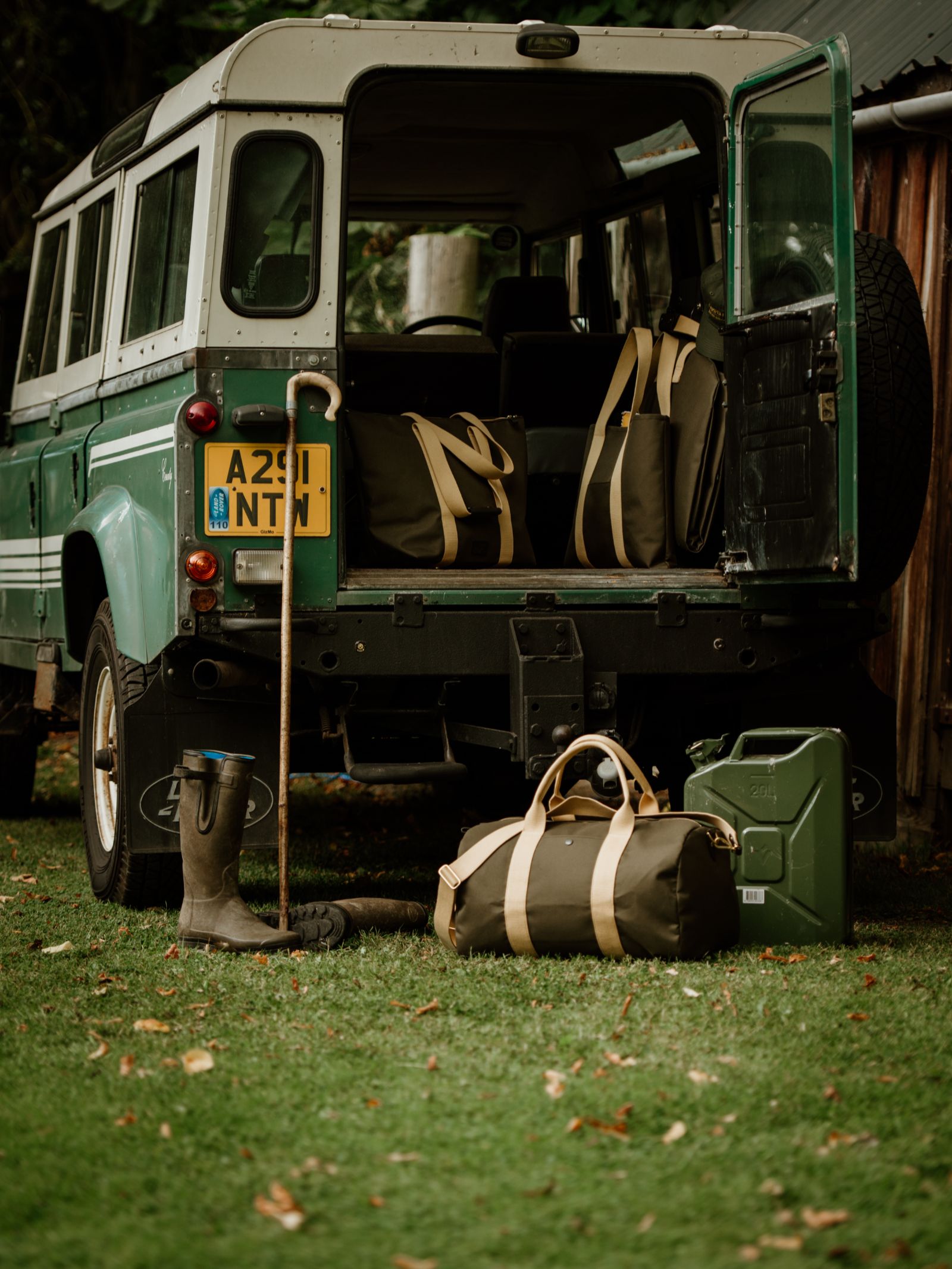 A green Land Rover with its rear door open reveals bags. On the grass behind it are a Campbell's of Beauly Utility Medium Duffle, a jerry can, wellington boots, and a walking stick, set against trees and a wooden structure.