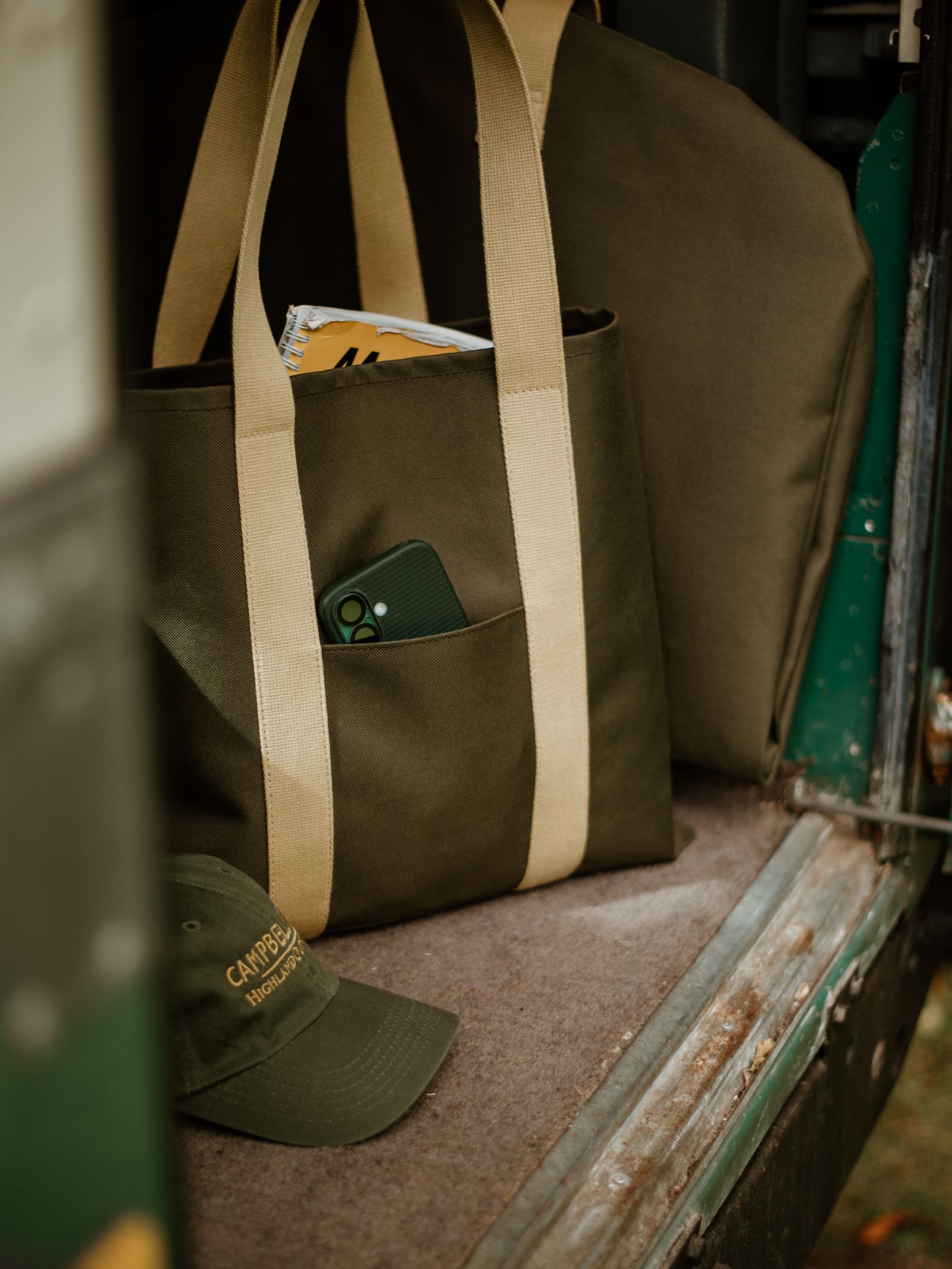 The Campbell's of Beauly Utility Packaway Tote in olive green canvas with beige handles is shown on a vehicle, holding a green phone in its pocket and a magazine inside. A green Camp Gear hat is placed nearby.