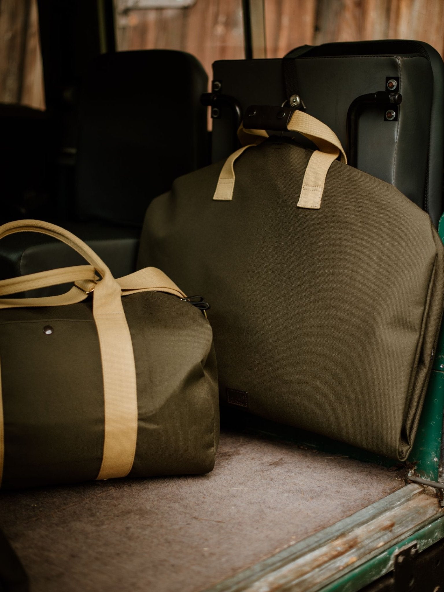 Two Campbell's of Beauly Utility Suit Bags in olive green heavy-duty canvas with tan handles are displayed on a vehicle seat—one upright, one flat—offering durable garment protection. A wooden building is visible in the background.