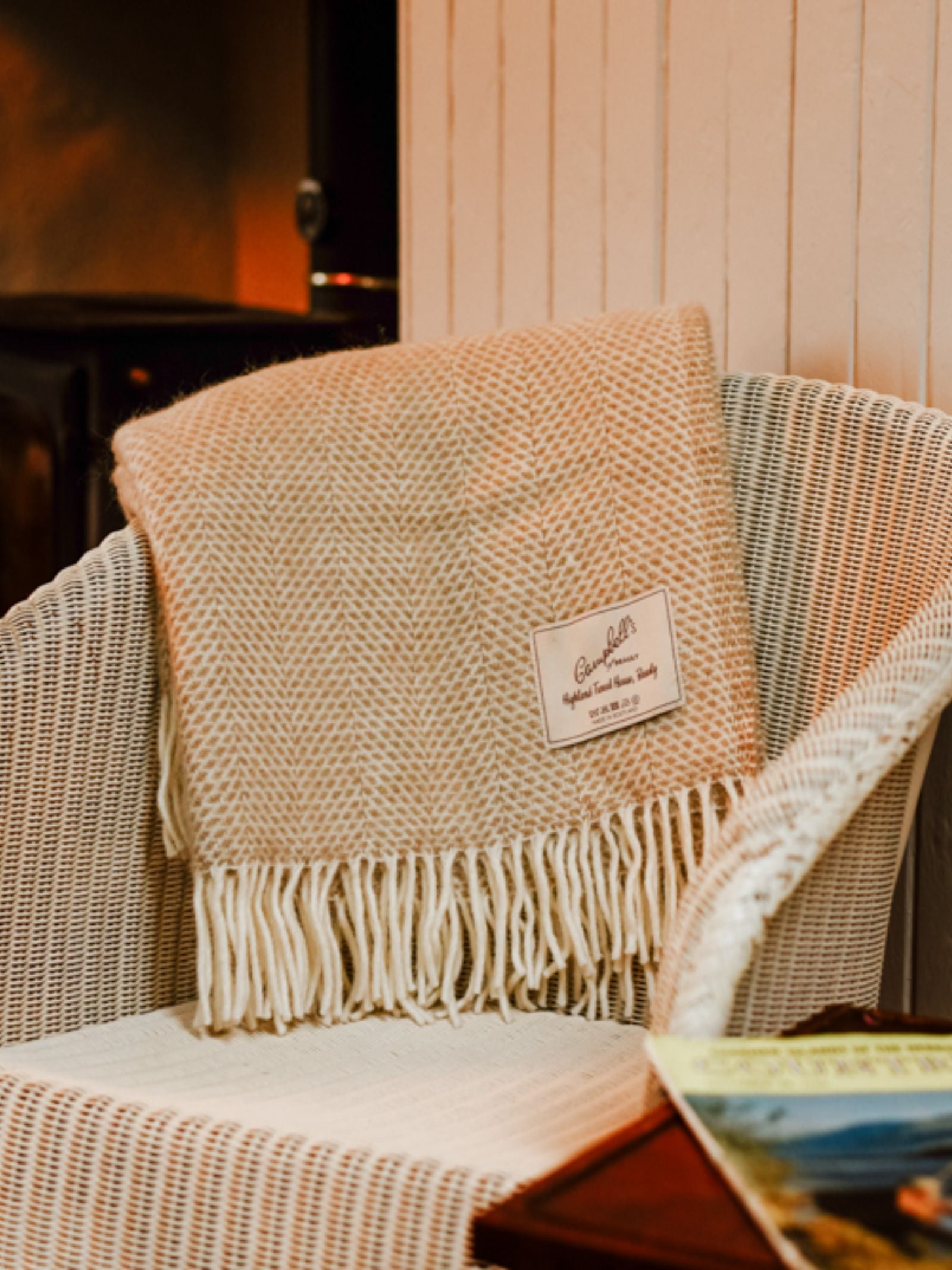 A Campbell's of Beauly Stipled Twill Blanket in cream-colored wool, featuring a breathable weave and fringe, drapes over a white wicker chair with its label visible. A magazine sits on the nearby table.
