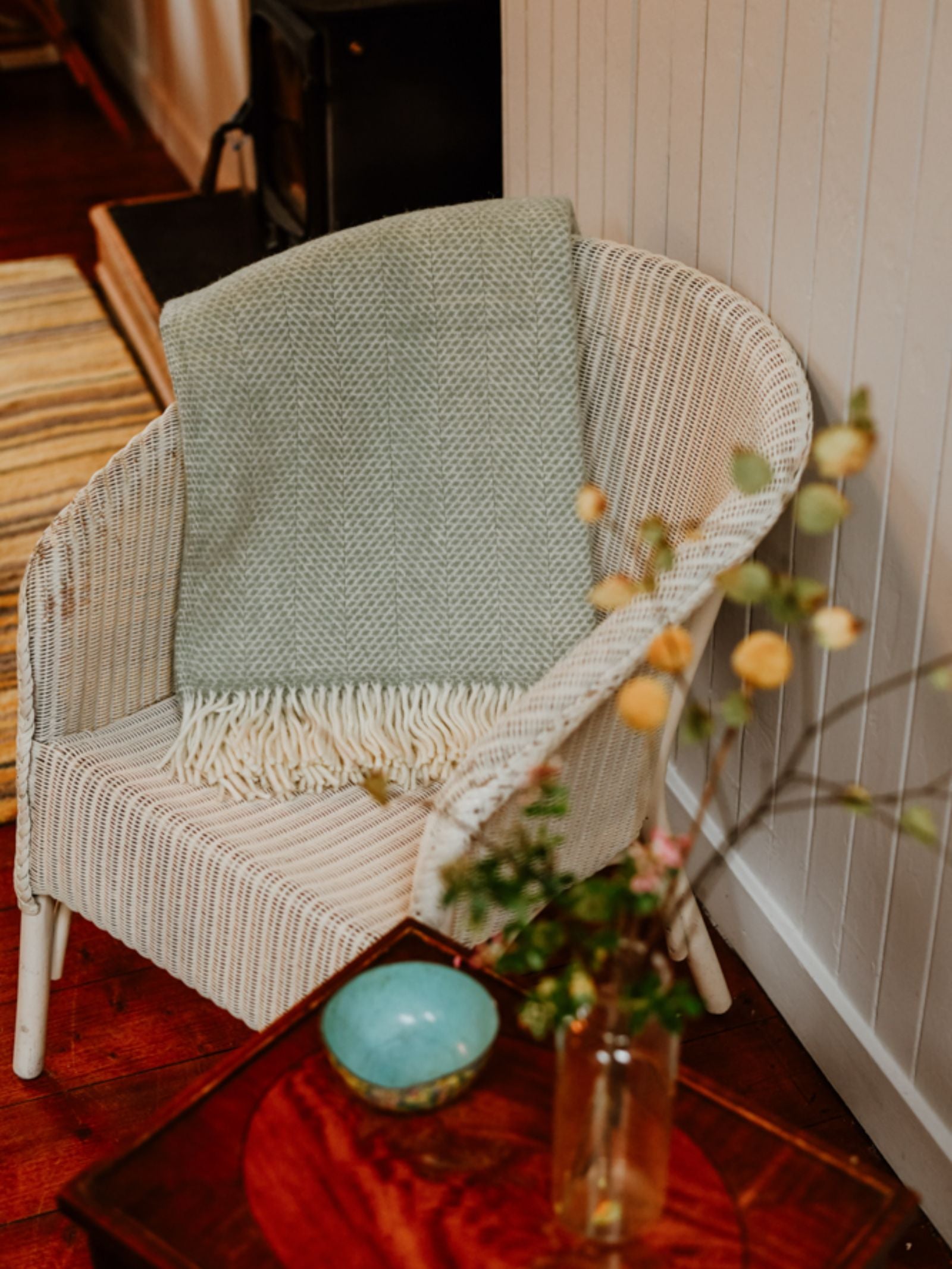 A white wicker chair with a Campbell's of Beauly Stipled Twill Blanket draped over it sits beside a wooden table holding a small blue bowl and a vase of yellow flowers, creating a cozy indoor setting.