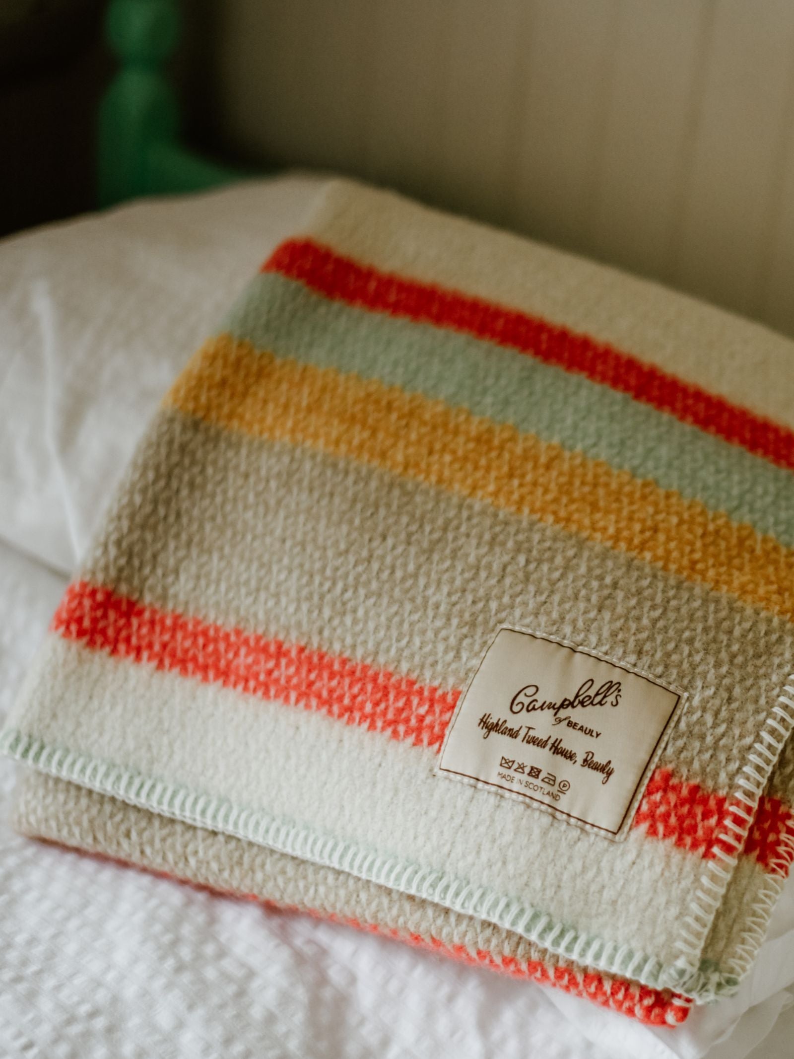 A folded wool Baby Blanket by Campbell's of Beauly, featuring red, yellow, and green stripes, lies on a white bed. The cozy and inviting setting highlights its traditional design and quality label.