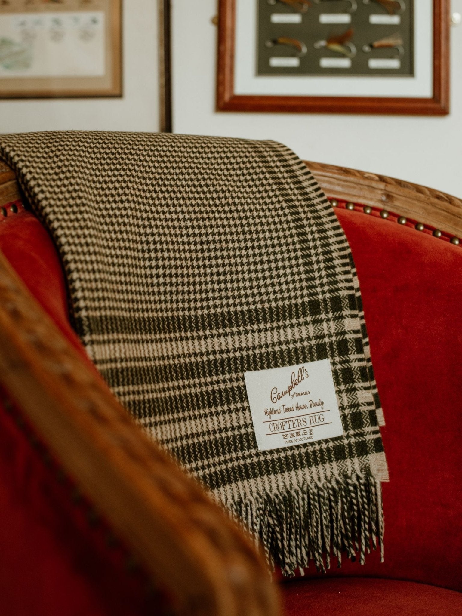A black and white houndstooth Crofters Blanket by Campbell's of Beauly, with fringed edges, is draped on the back of a red velvet armchair for a cozy country look. The branded label is visible. Framed art hangs on the wall.