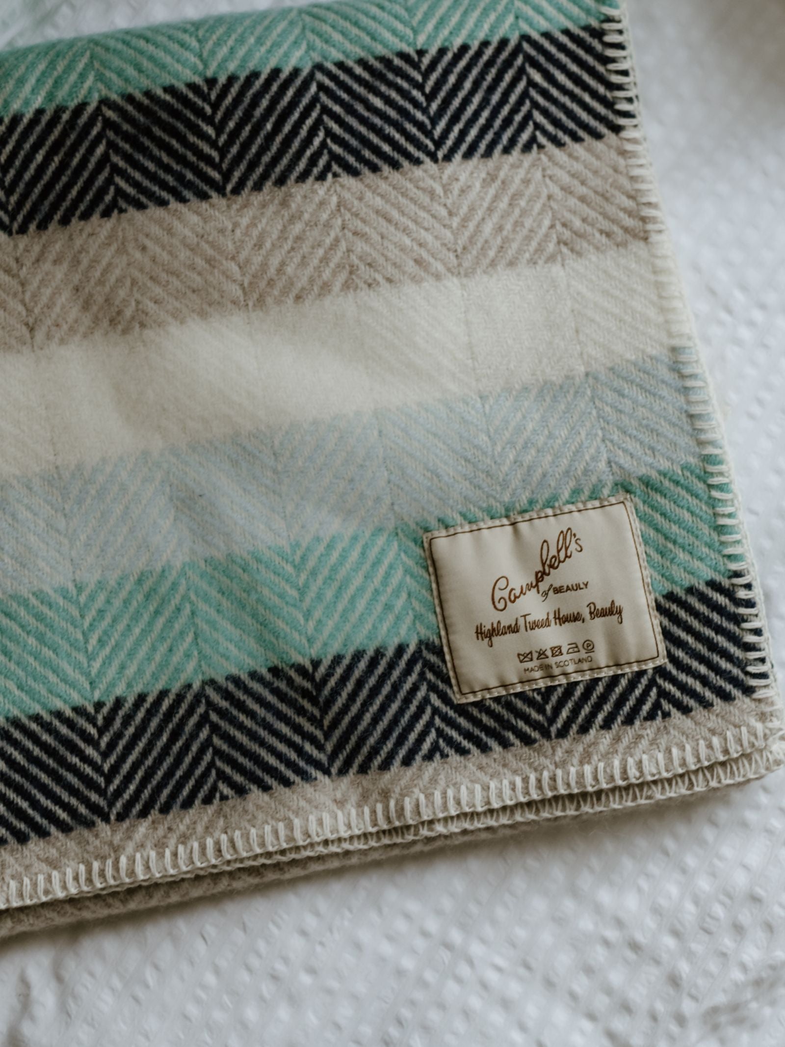 A close-up of the Campbell's of Beauly Baby Blanket, a lightweight wool pram blanket with a chevron pattern in beige, blue, green, and black, resting on a textured white surface.
