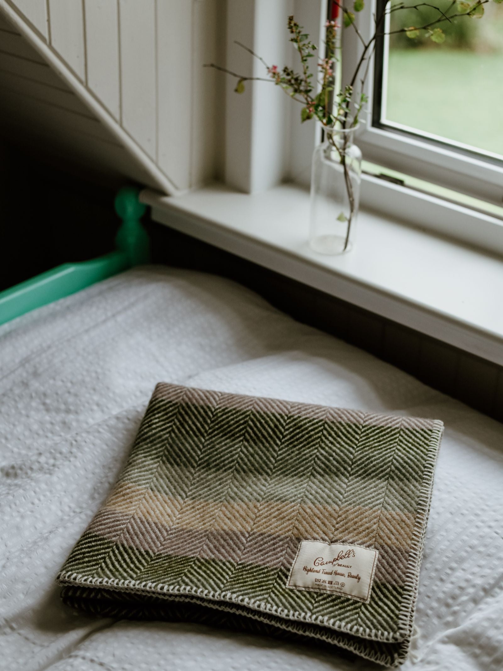 A Campbell's of Beauly Baby Blanket—folded, green and beige with a herringbone pattern and label—rests on a white textured bed near a window, where a glass vase with small branches sits on the windowsill.