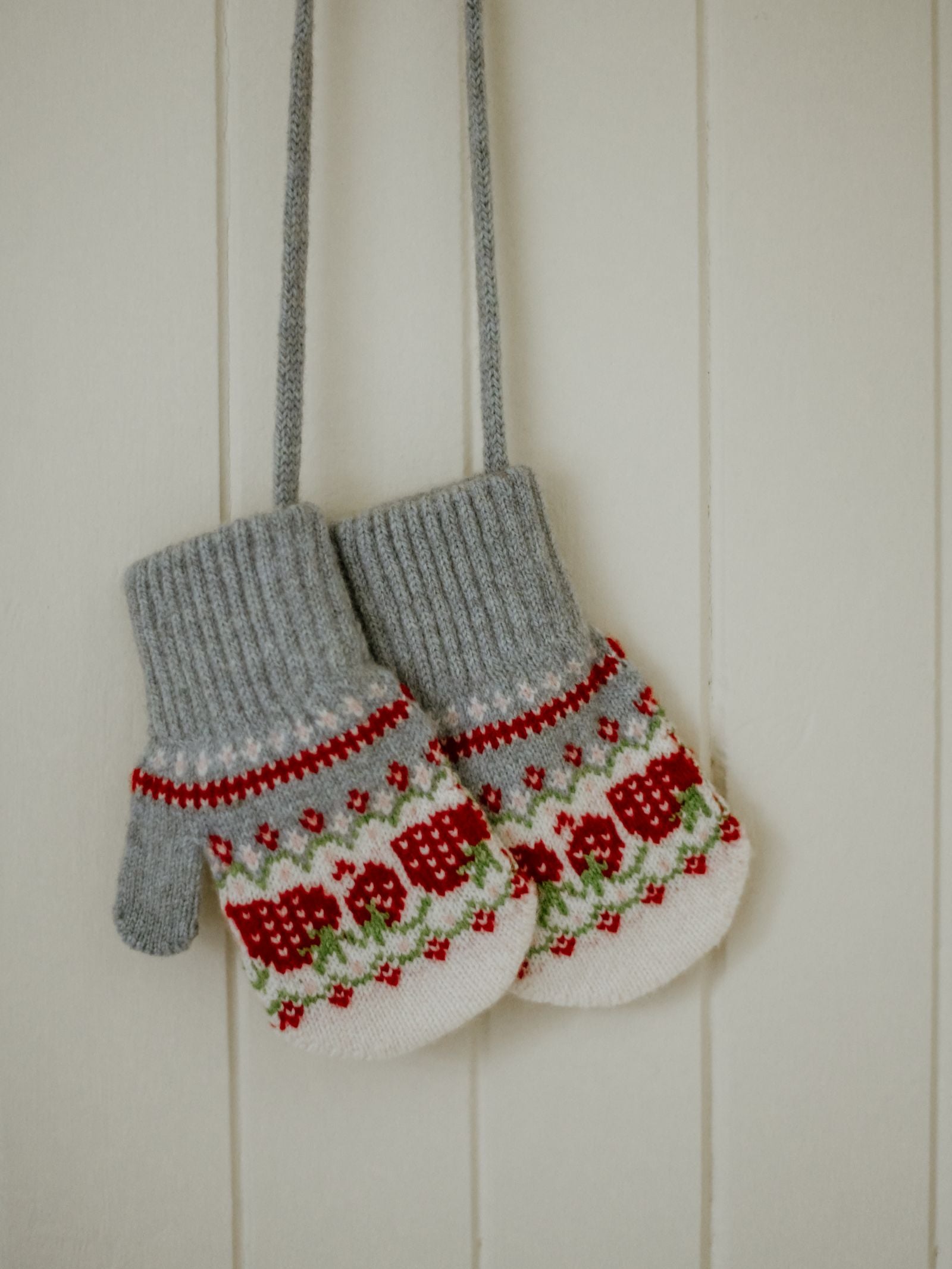 A pair of Campbell's of Beauly Mini Lambswool Strawberry Mittens with gray cuffs and a red, green, and white pattern hang by their string against a white wooden wall.